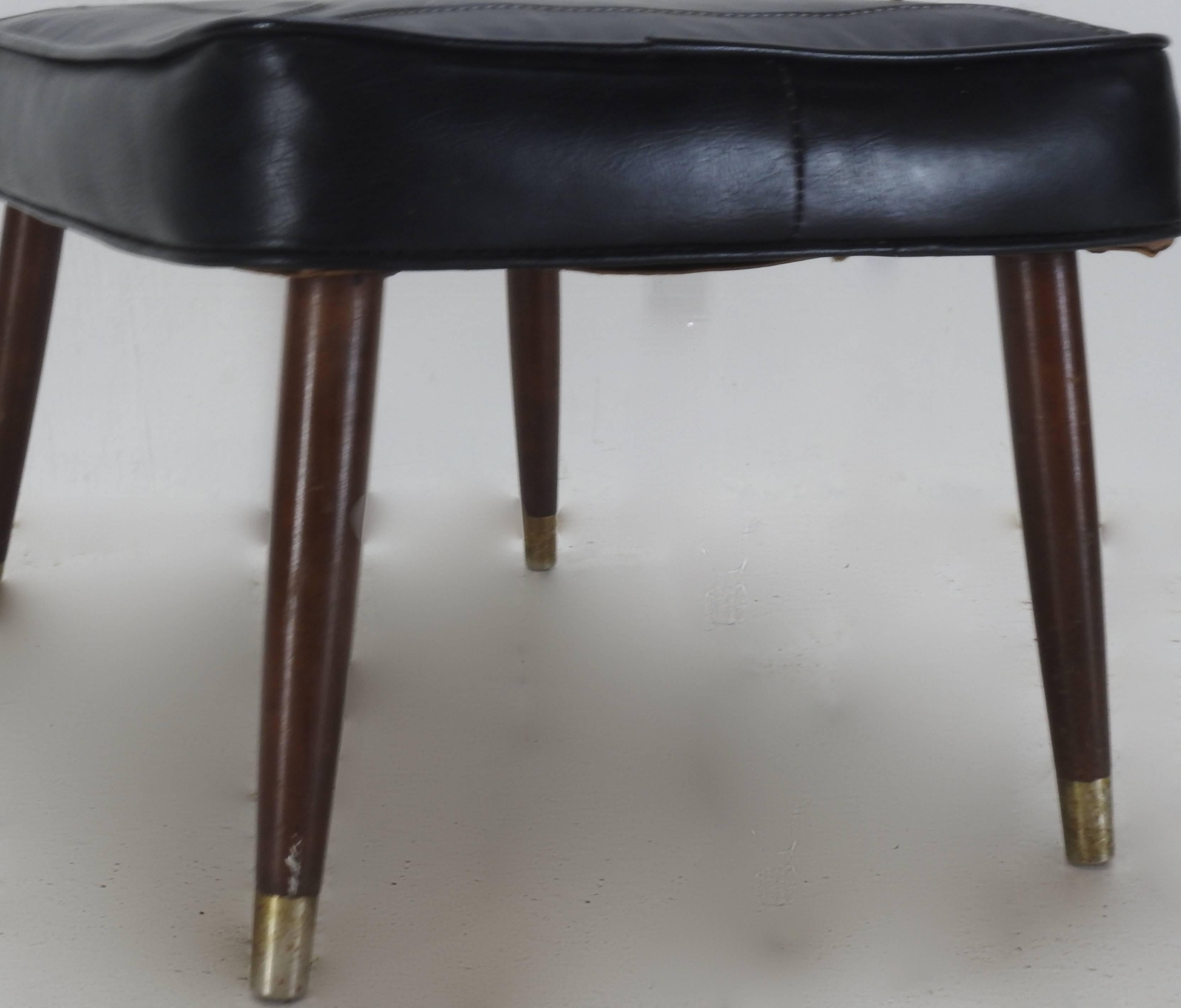 This is a Classic Mid-Century Modern footstool made up of black leather and wooden legs capped off with brass. The stitching adds to the looks of this useful stool.