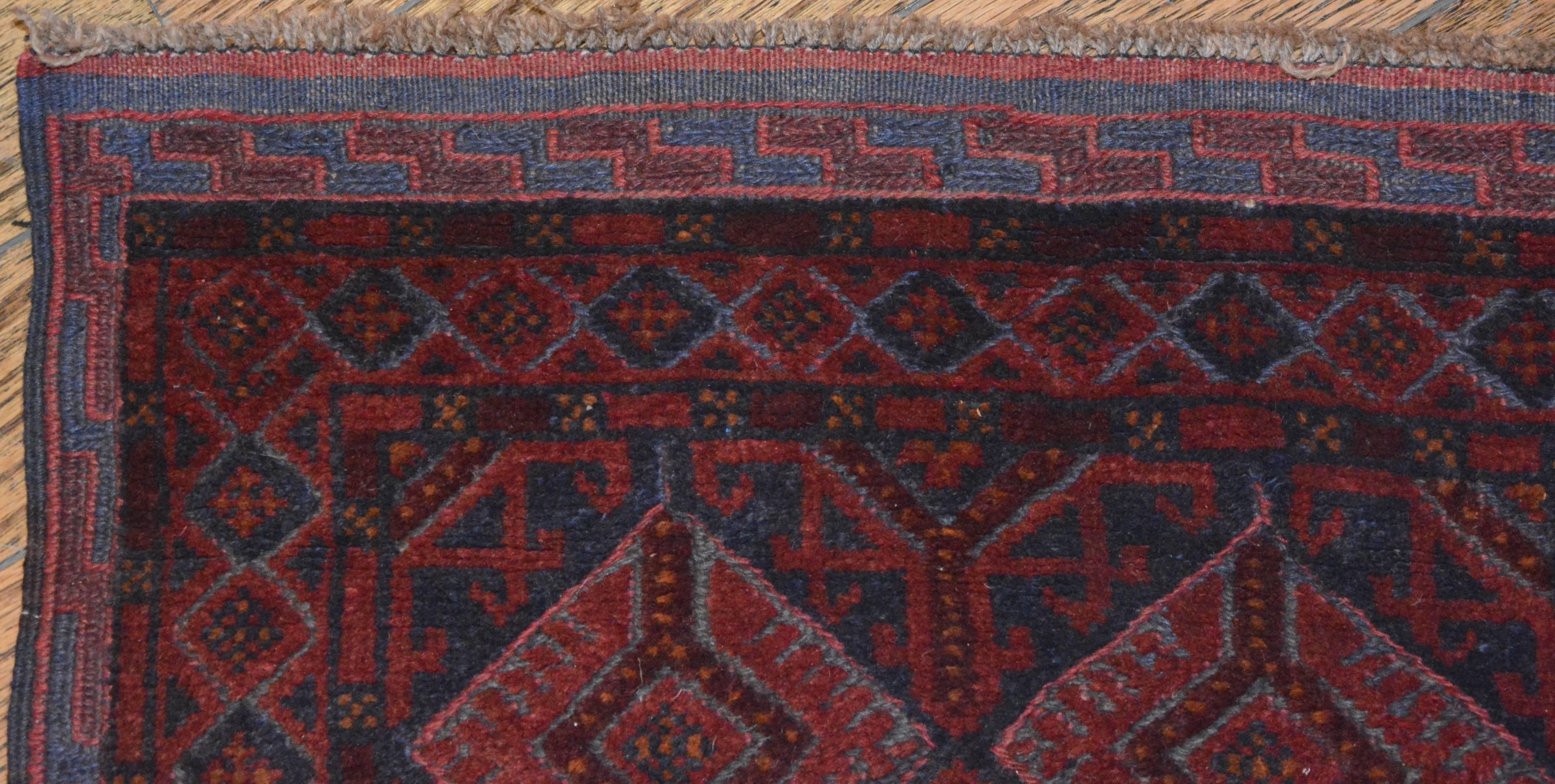 A hand knotted and woven Baluch tribal runner. This geometric wool rug is rendered in a palette of red, burgundy and midnight blue. It features an all-over pattern of latticed Tekke-style diamond medallions and Kurbage guls over a midnight blue