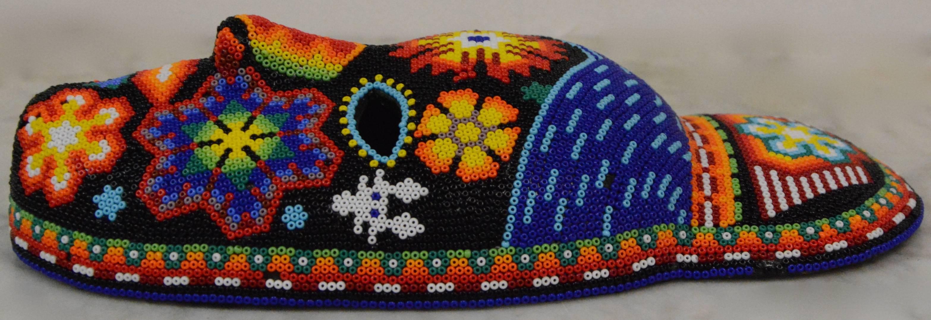 This is a colorful hand beaded Huichol Folk Art mask. The mask features intricate tribal-like designs throughout that are comprised of glass beads in vibrant shades of red, orange, yellow, green, blue, purple, black, and white. The beads are mounted