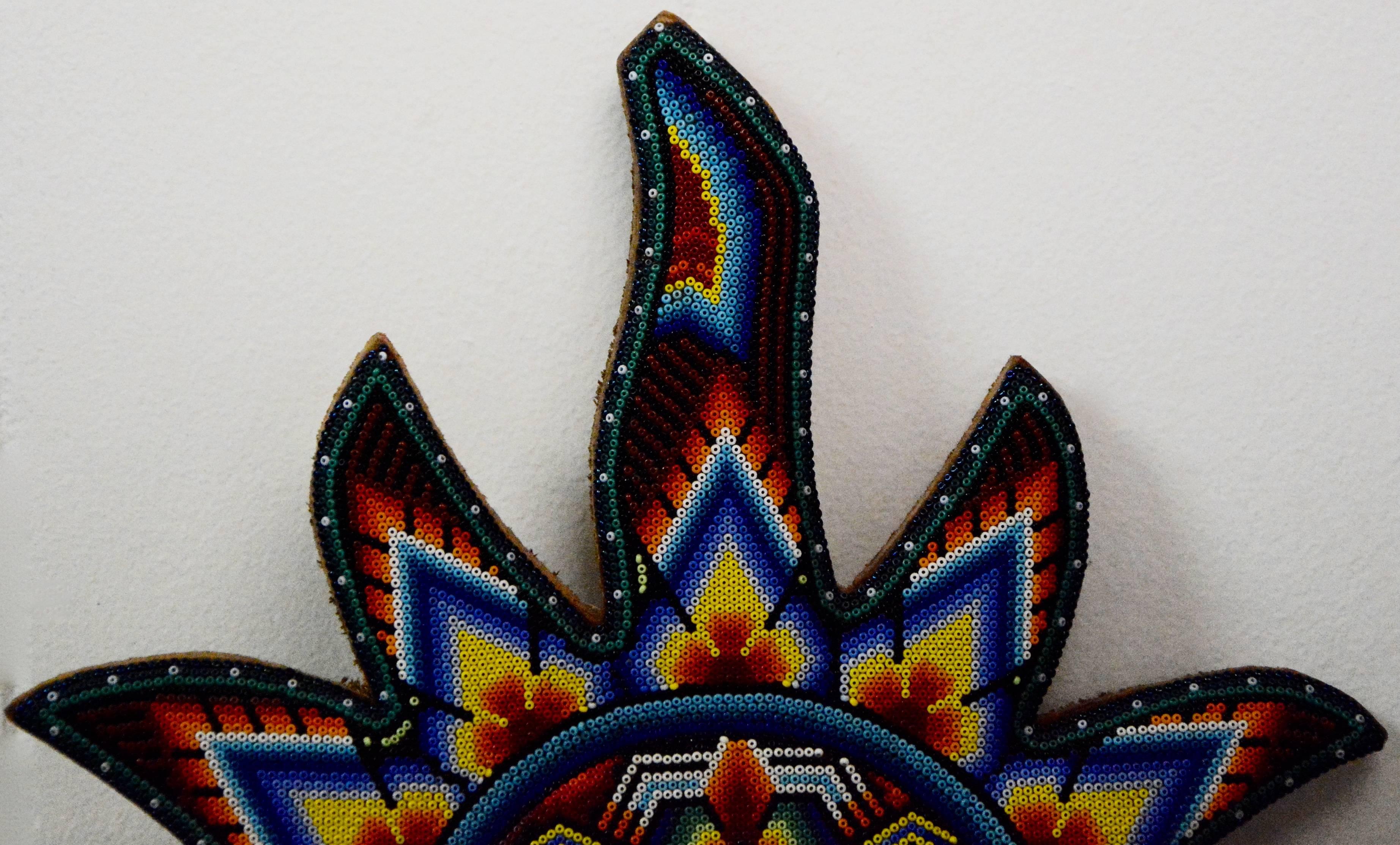 This is a very intricate hand beaded Mexican Huichol sun wall decor. This unique piece is comprised of vibrant glass beads in shades of red, yellow, orange, green, blue, grey, black, and white, which are arranged in the shape of a sun and mounted on