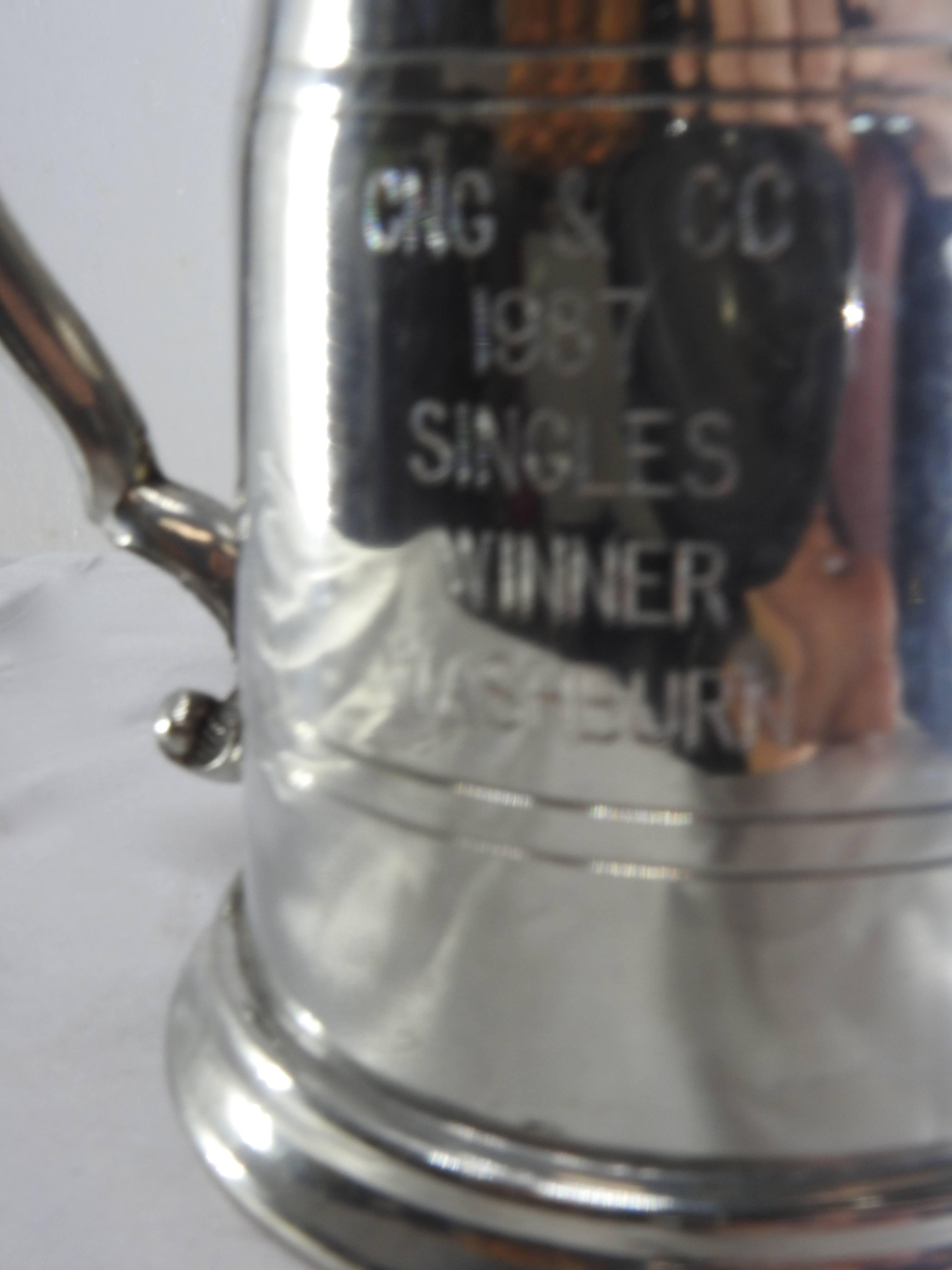 This Sheffield tankard is accented by a scrolling handle on the side. The tankard itself has curvy lines to add to its beauty. It in engraved CNG & Co., 1987, Singles Winner, Washburn. Pewter forms the tankard and the bottom has clear glass. The