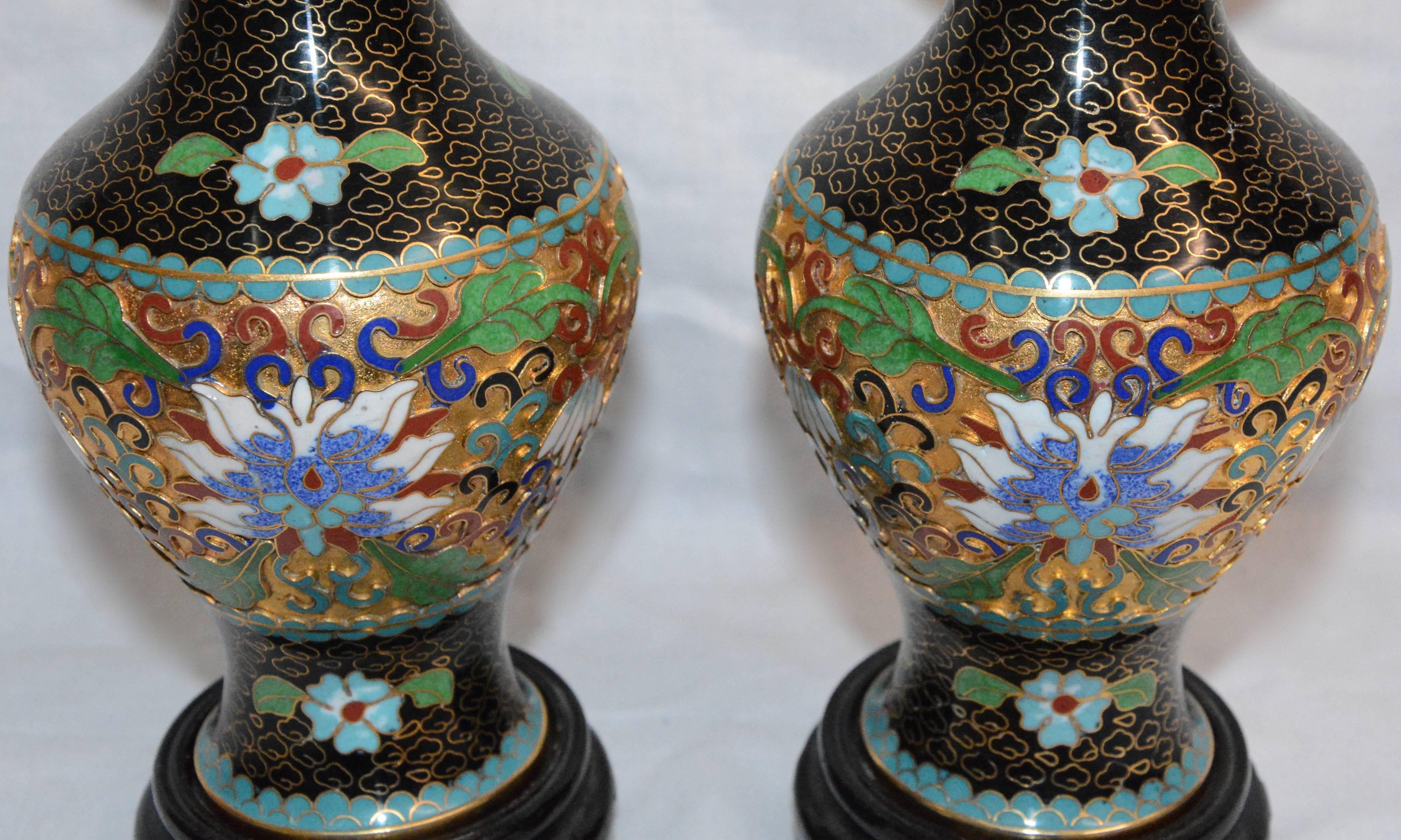 This is a colorful pair of Chinese cloisonné vases that sit atop black lacquered wooden bases. Beautiful flowers encircle the vases and are adorned with stunning details around the vases. The tops have a gold finish. The set includes the original