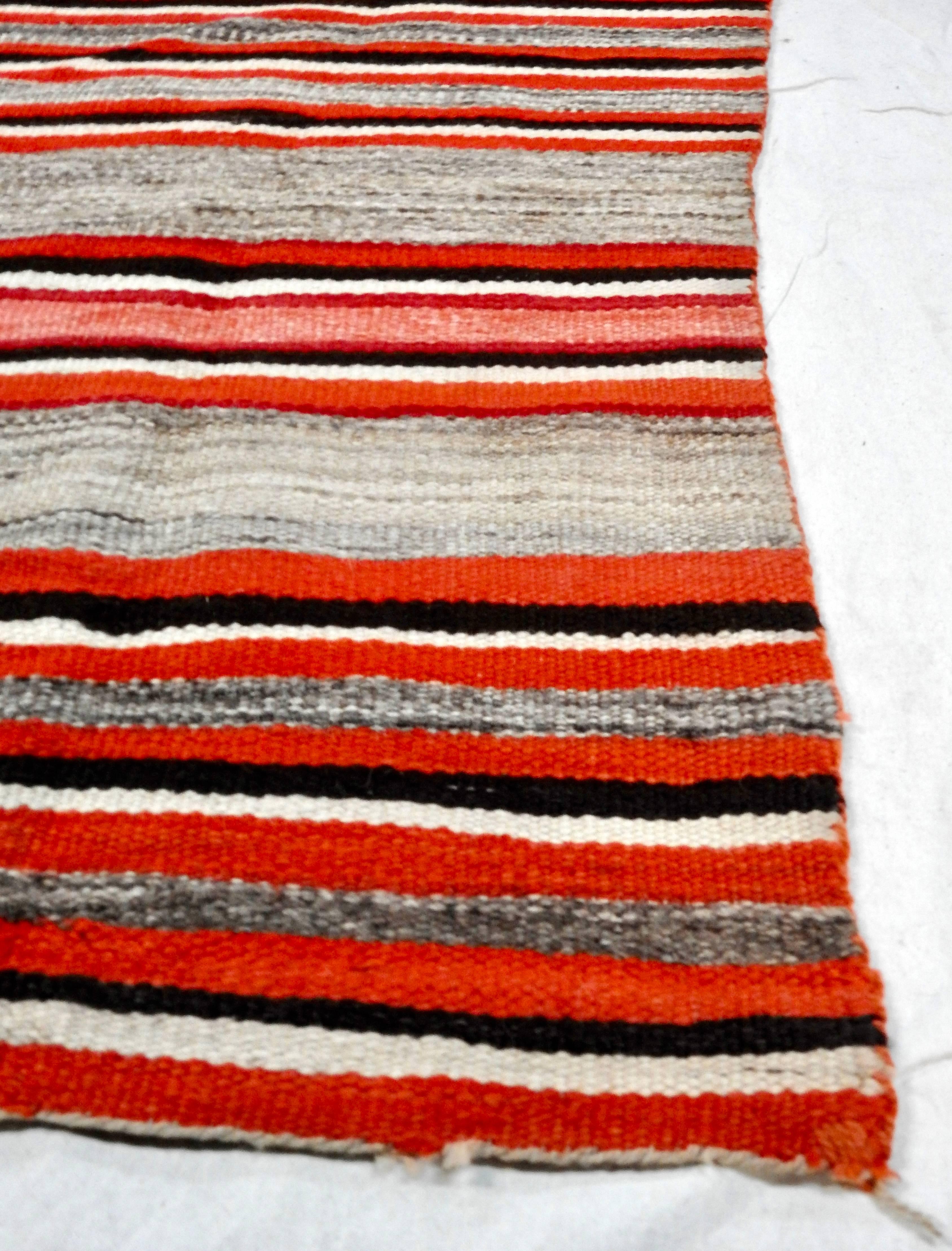 Featured is an antique, circa 1890s handwoven Navajo transitional wool rug. The rug features bold stripes of various widths, in a palette of red-orange, cream, tan and black, with tan and brown twisted stitches to the edges, and short twisted