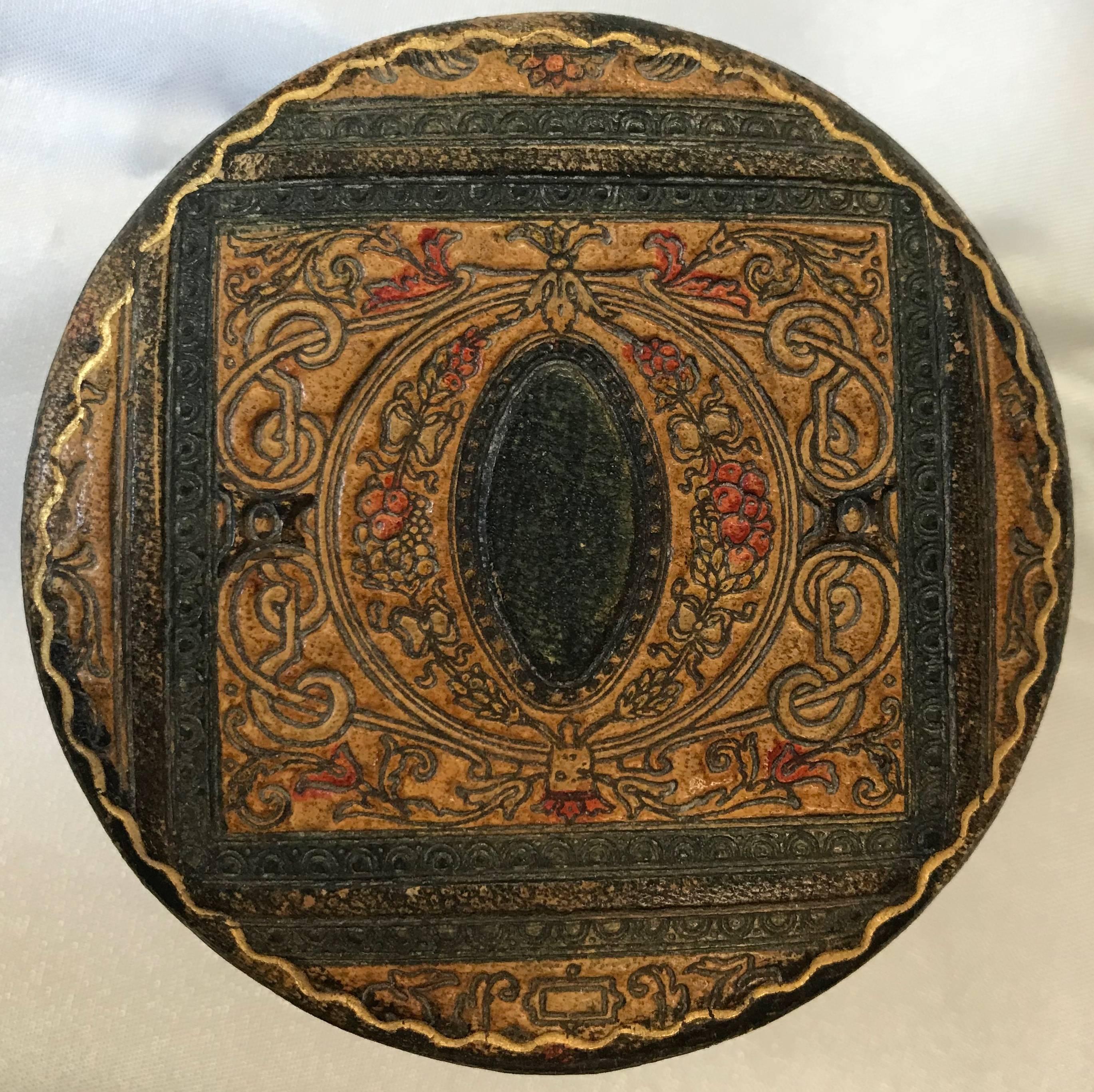 Embossed leather tinted black brown, red and gold is displayed beautifully on this round box with a medallion on top. The lid lifts off to a lining of black satin.