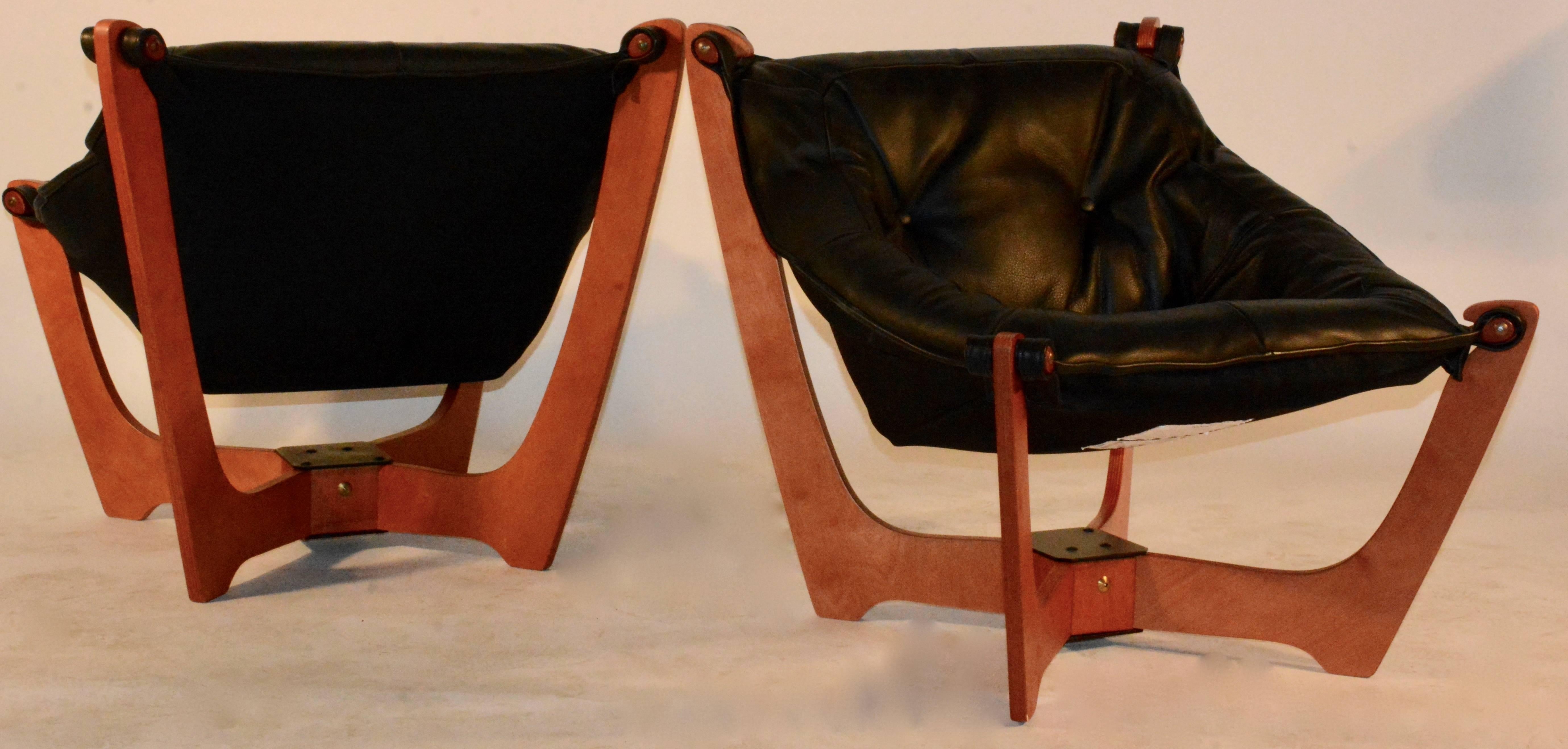 This pair of Luna sling chairs are made of birch plywood with a warm natural tone. The four legs come out of a square base on an angle with soft curves leading upward. The seating is covered in black leather and attaches to the legs with a knob like