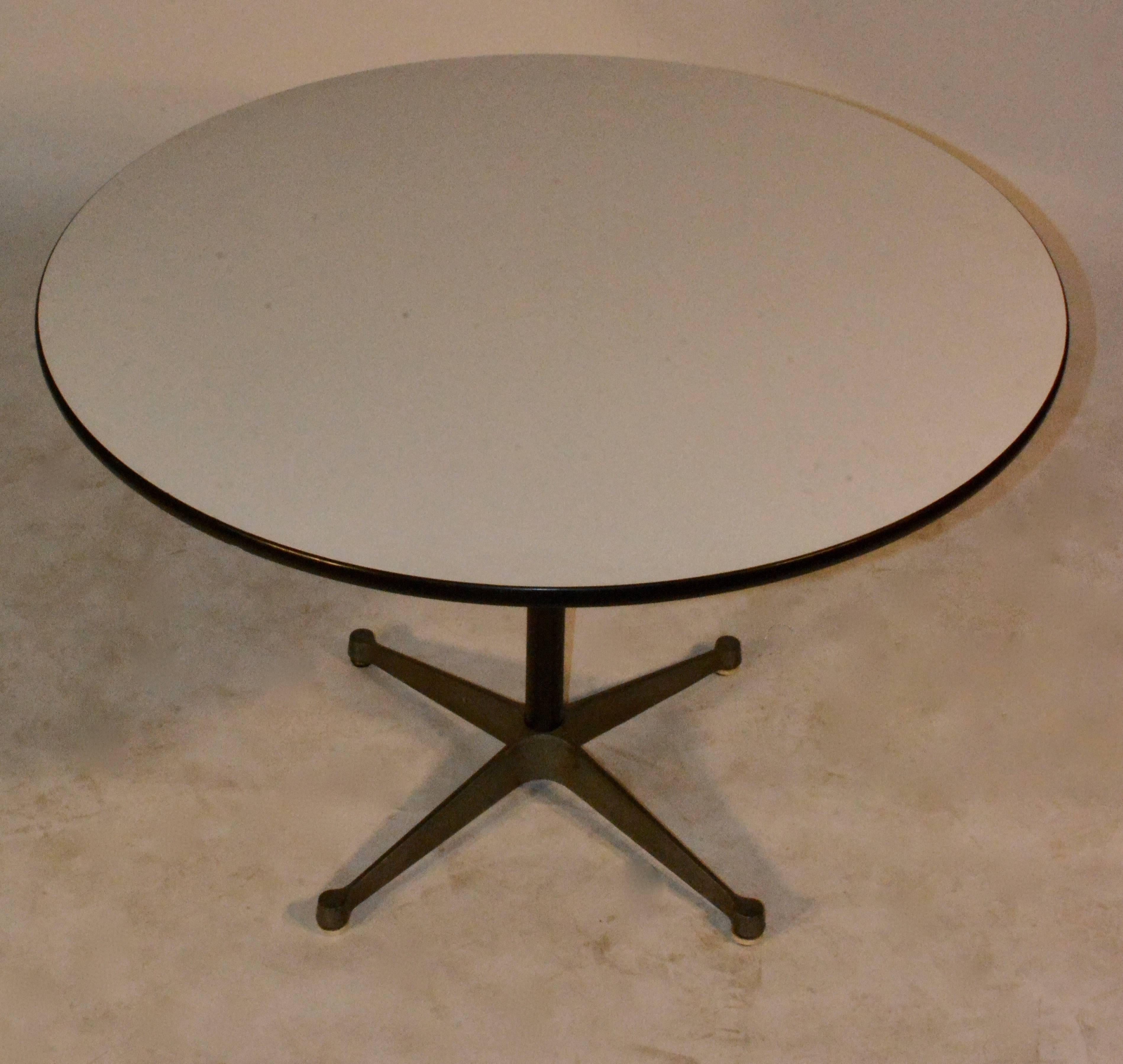 A midcentury Eames Aluminium group dining table by Herman Miller. The table features a round, white laminate top above a cylindrical, steel support ending in a four-point, aluminium base. The underside of the top retains a sticker for “Herman