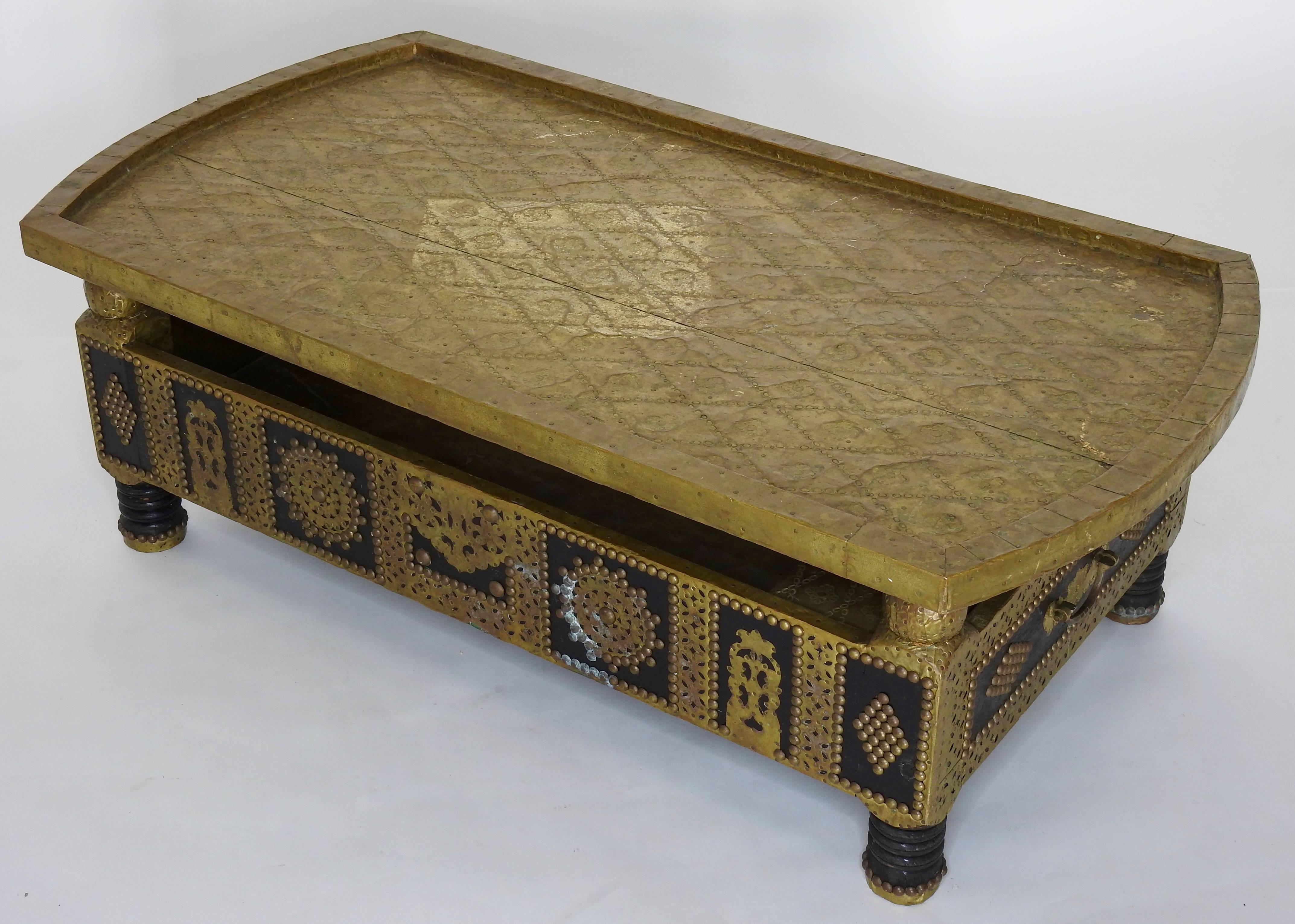 This is an ornate wooden coffee table detailed with hammered brass and tacks. The top can easily be removed for storage. The brass boasts a nice aged patina. The table is signed but we cannot determine the maker.