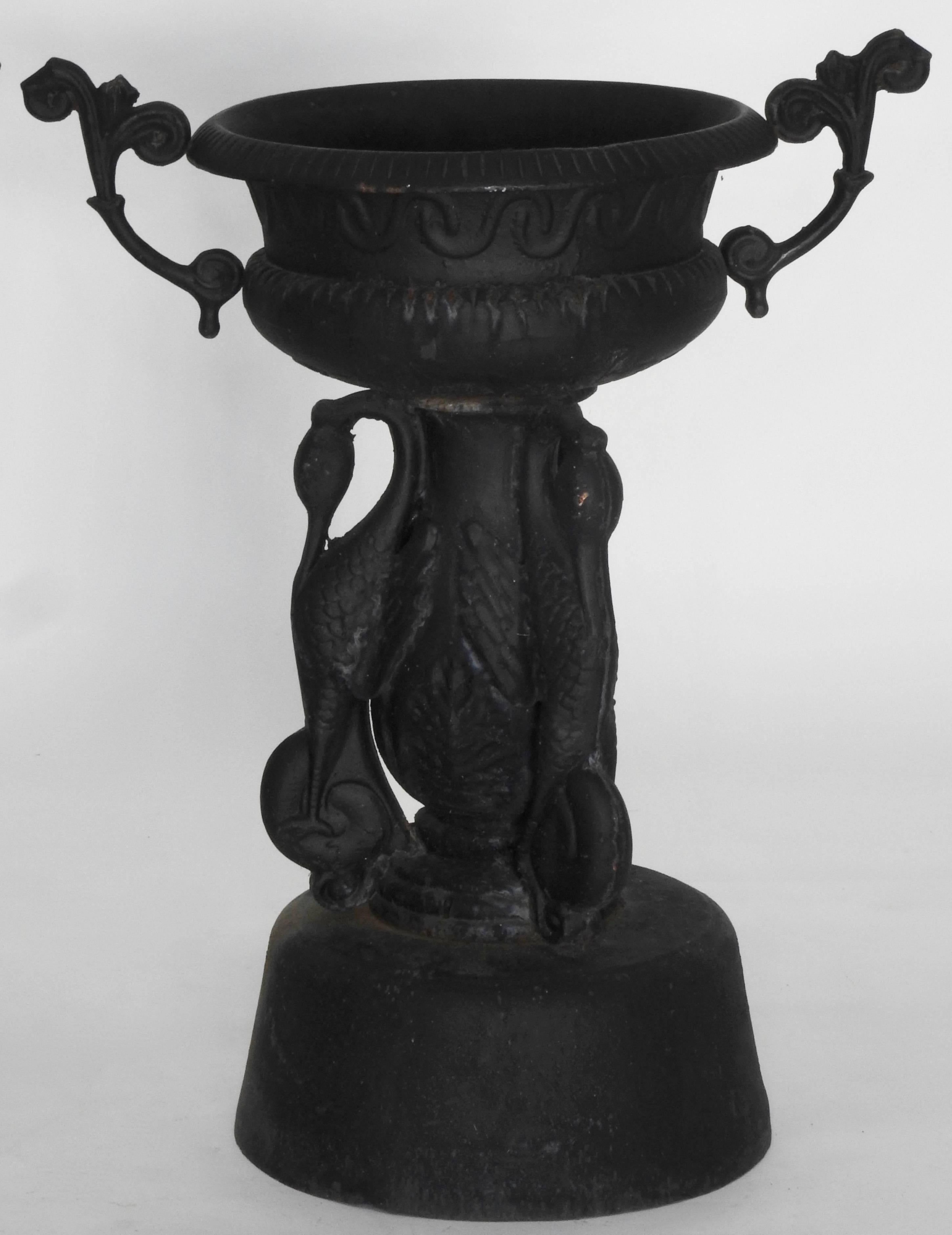 This is a spectacular pair of matching cast iron garden urns featuring cranes and ornate handles. They rise on a round riser followed by the cranes that rise with feather details and the rounding of the neck. The vessels sit atop the cranes with a