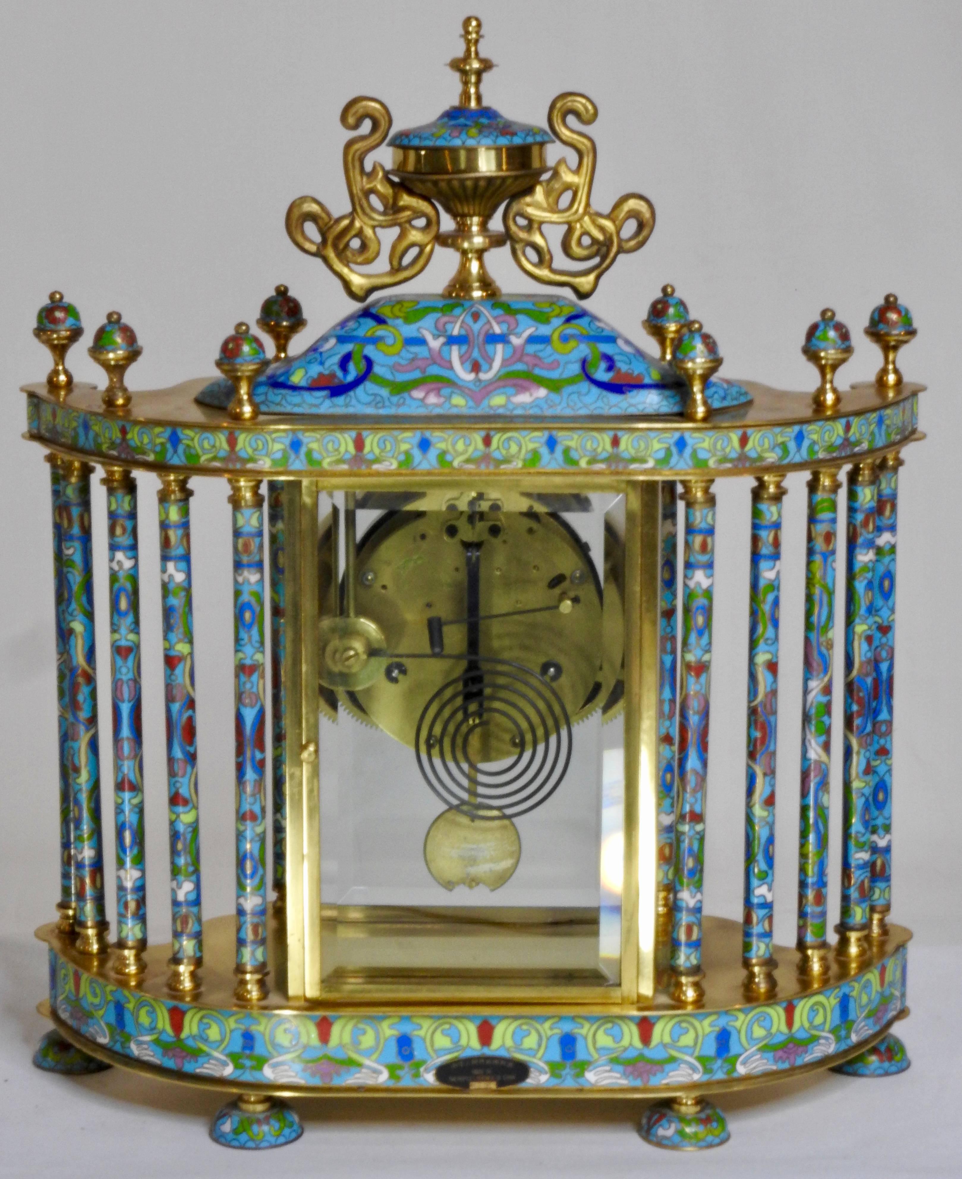 An antique Chinese shelf clock. It is bronze inset with cloisonné. The clock face is decorated with flowers and has black Roman numerals. There is a glass door that opens to a gold tone pendulum. There are ten columns decorated with cloisonné̀ and