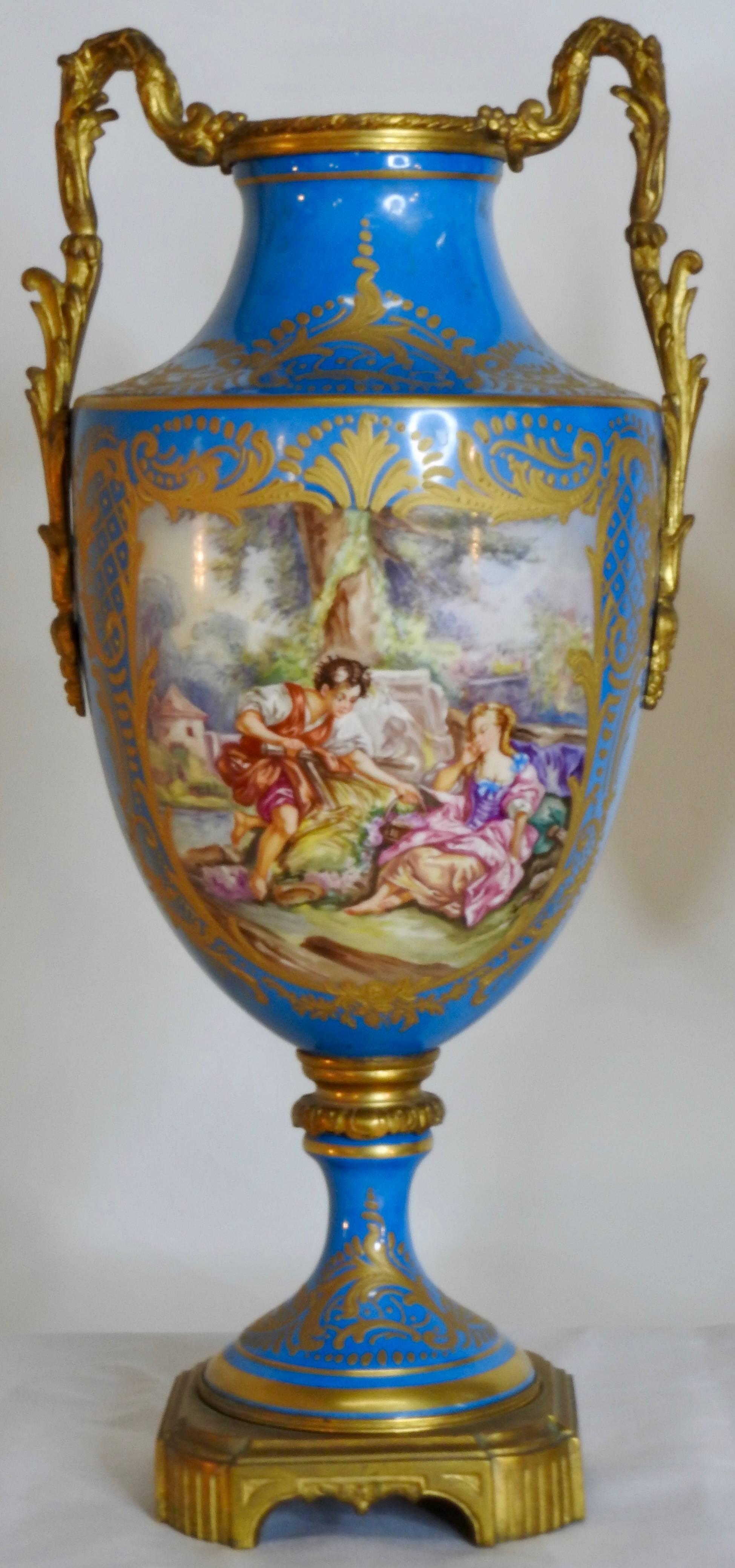 This gorgeous pair of sevre urns start at the bottom with a square bronze gilded base. Rising to the porcelain of the vibrant blue with gold paint detail to a neck piece of gilded bronze. Coming to the body of porcelain with different scenes painted