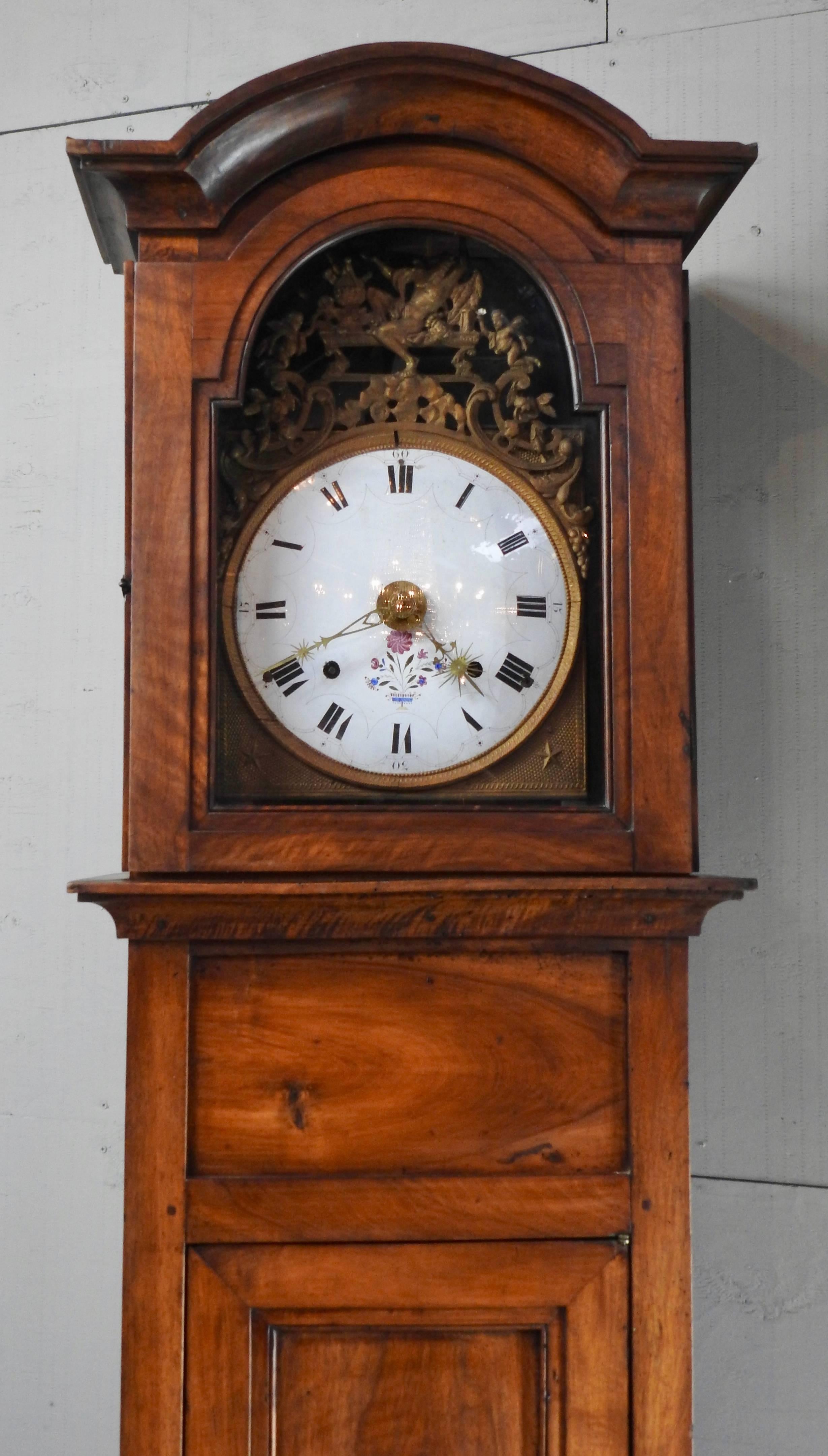 Cherrywood long case clock featuring hand-cut metal hands and Roman numerals. Dainty flowers add to its beauty. The bonnet has graceful curves and tops off a medley of dancing cherubs in gilt. There is a window in the door to view the bronze