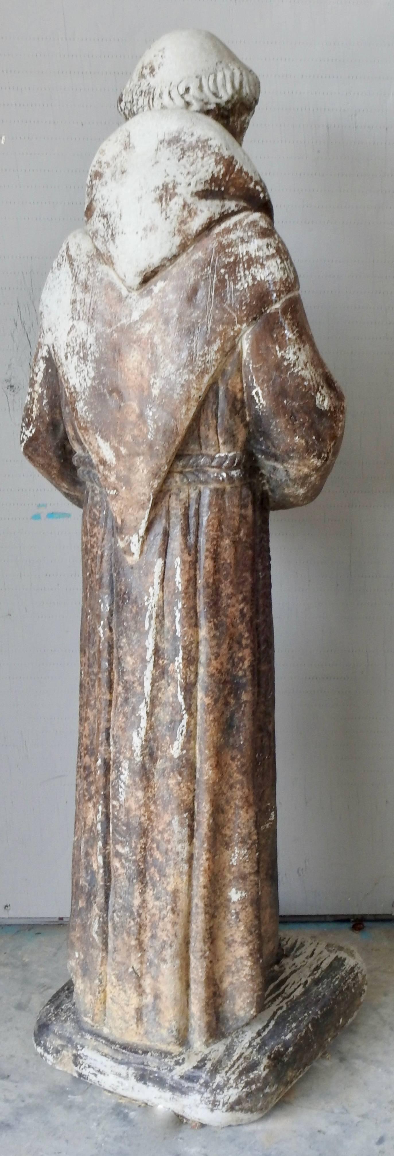 A cast stone garden statue of St. Francis of Assisi. The statue has been outside and has developed a weathered patina which adds to his character. The applied colors have faded over the years. He is holding a bird in his hands.
  