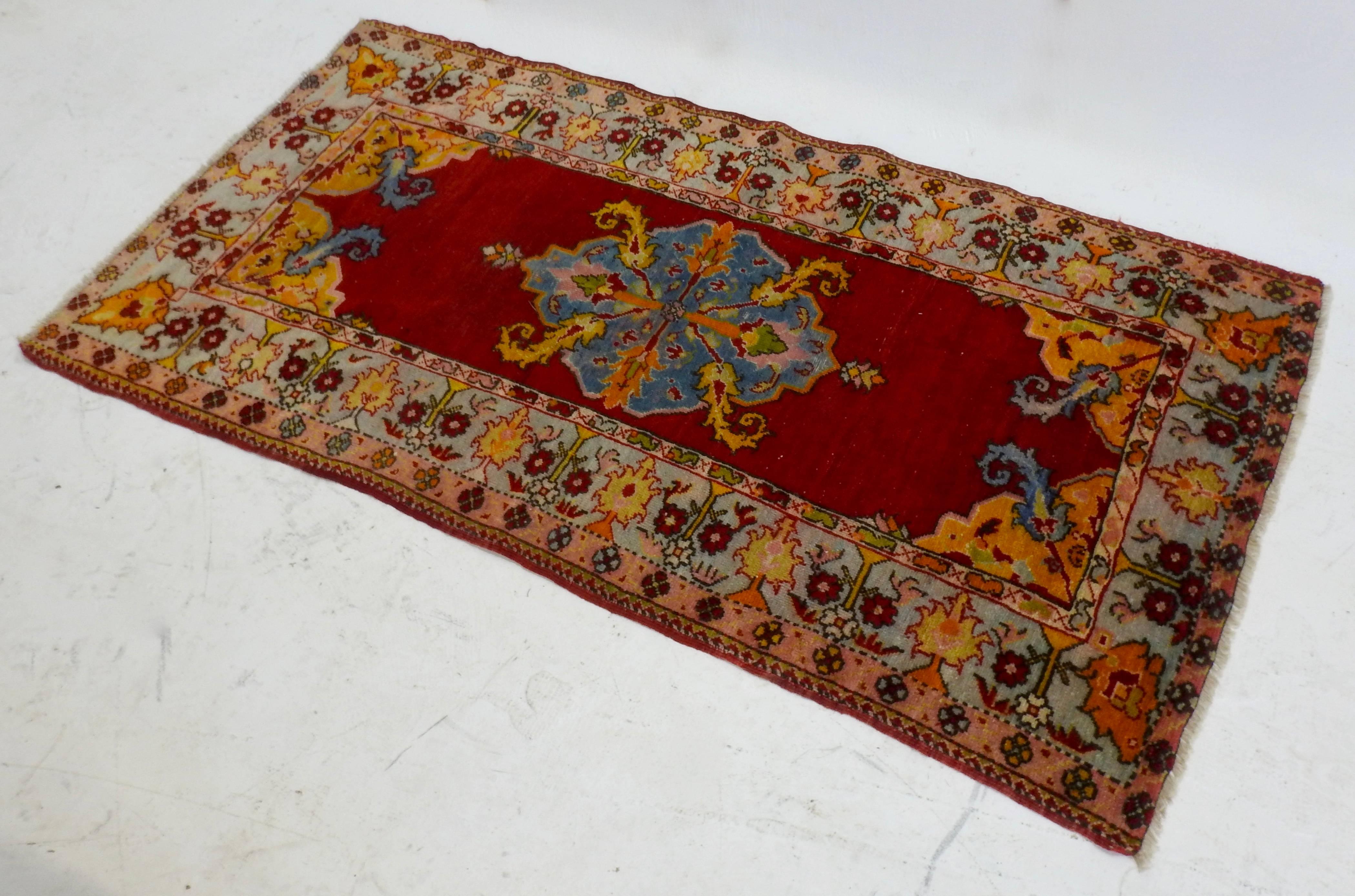 A brightly colored midcentury Turkish Oushak rug. The hand knotted rug is made of wool with a geometric design featuring a light blue medallion to the centre against a red field with a surrounding red, blue and yellow floral border. There is a short