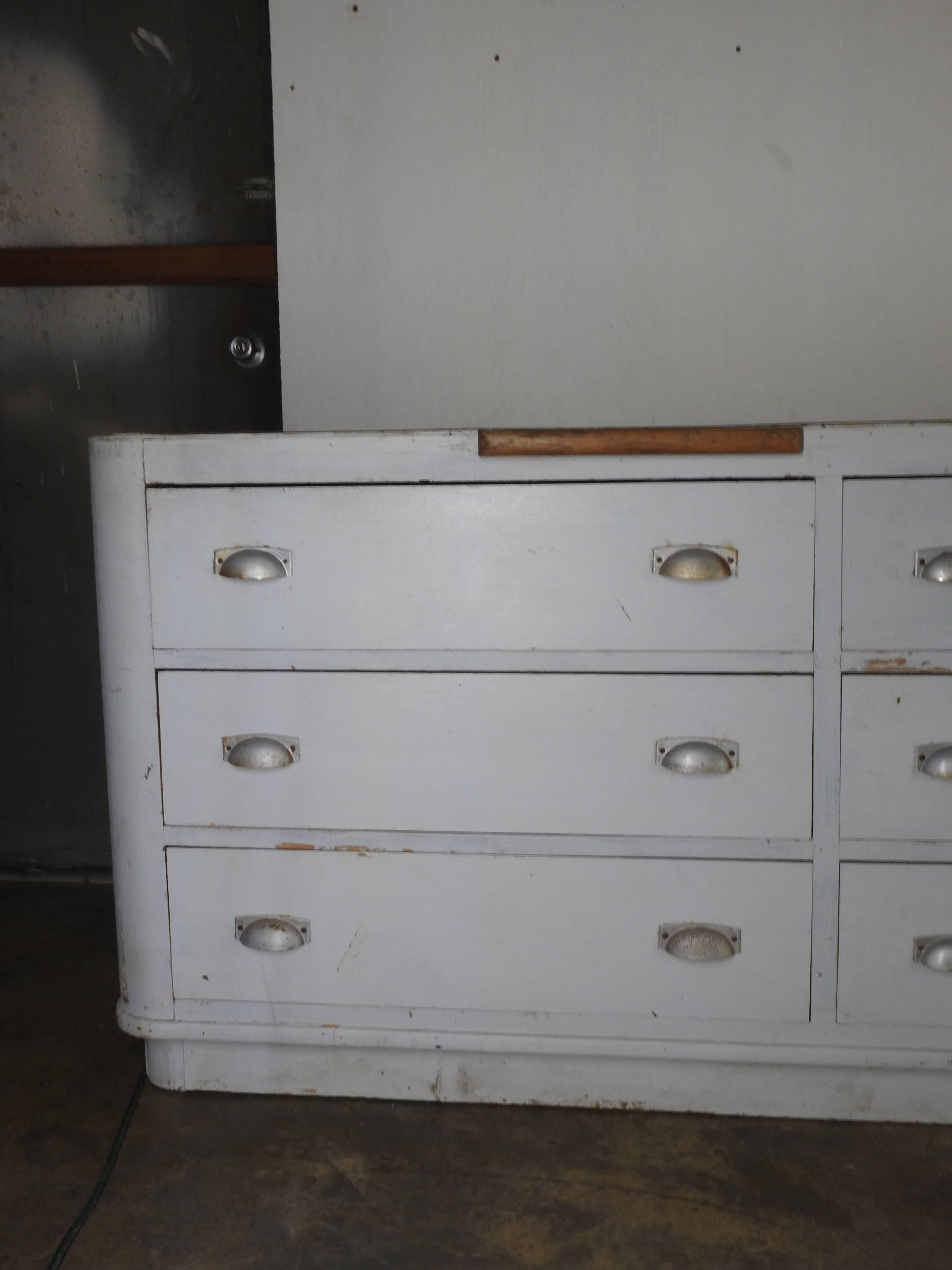 This is a French display counter and is painted in an off-white tone. It has nine large drawers for storage with an Industrial style pull in a painted silver finish. Also has to pull-out folding tables, or cutting boards.