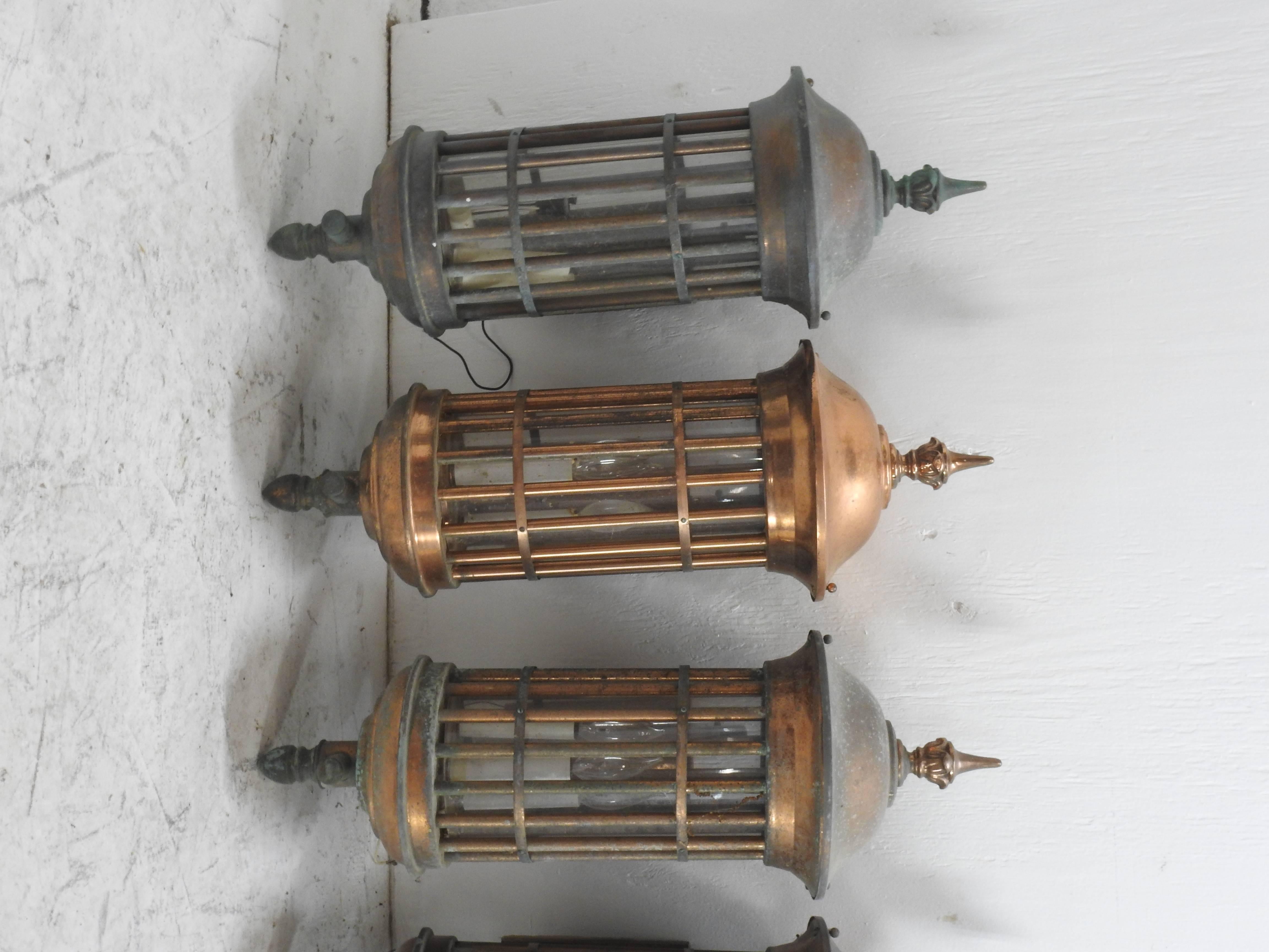 These are fabulous copper lantern style wall sconces. These lighting fixtures feature a cage like cylindrical body with a clear insert and three lights. The have slightly domed tops with foliate accented pointed finials. Each has a rounded finial