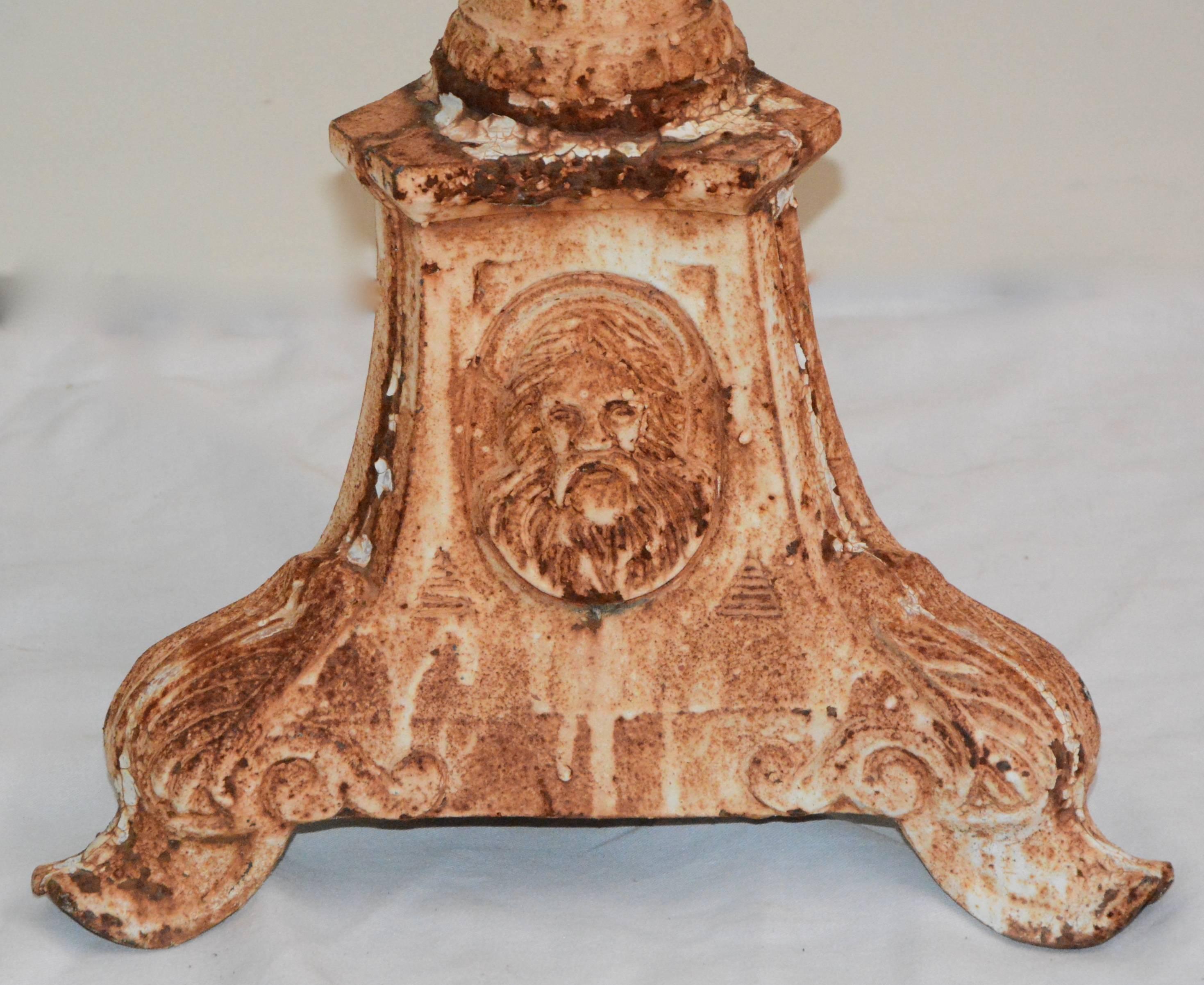 This is a substantial pair of cast iron candleholders. Each piece has a cast iron frame with white painted finish; featuring a single sconce with reeded detailing down the stem, a large base with an assortment of iconography vignettes to the sides