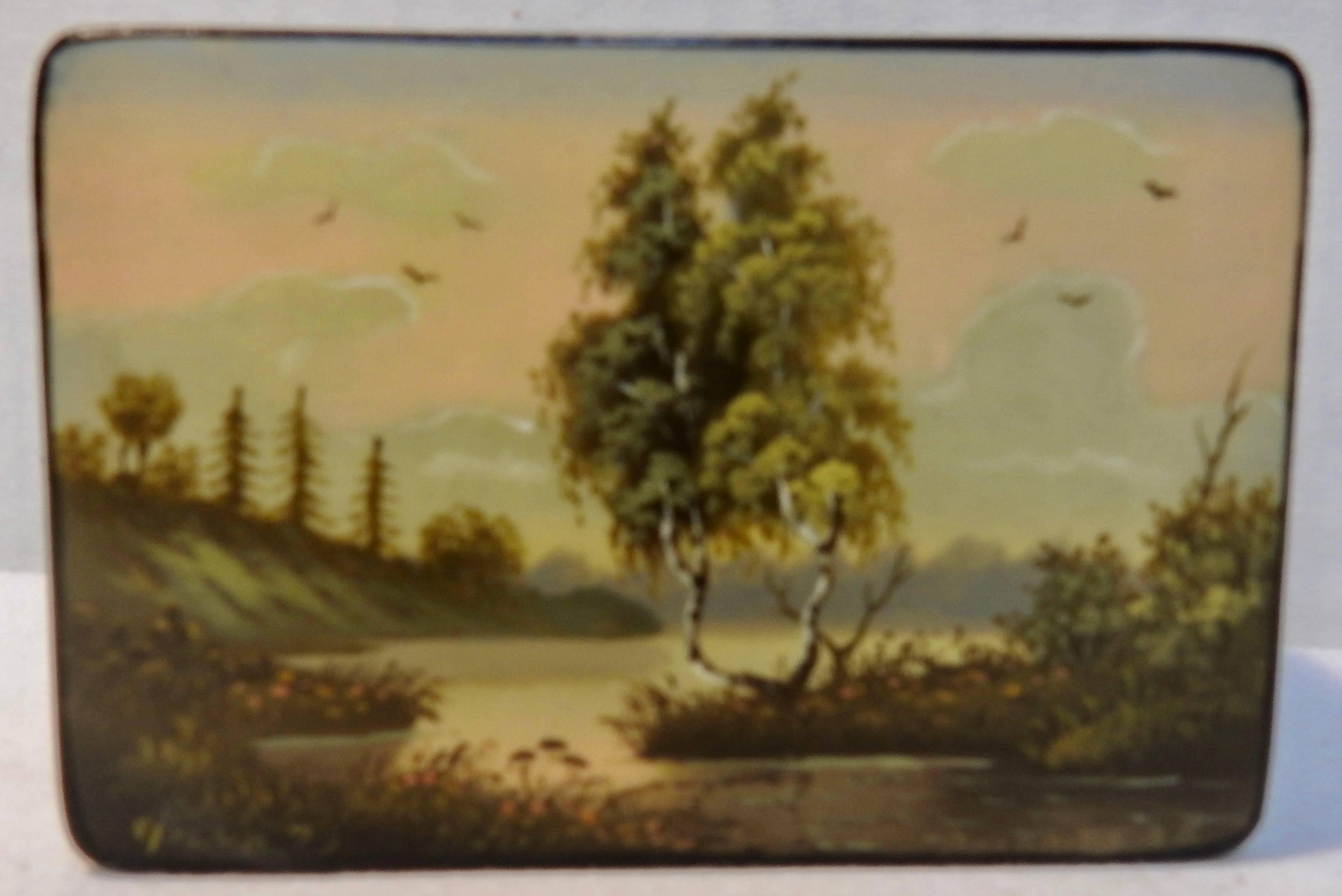 Featured is a Russian black lacquer snuff box with a vibrant red interior. The lid has a hand painted scene of trees along a river bank with birds flying around. The sky has soft, pastel colors. Dated 1973.