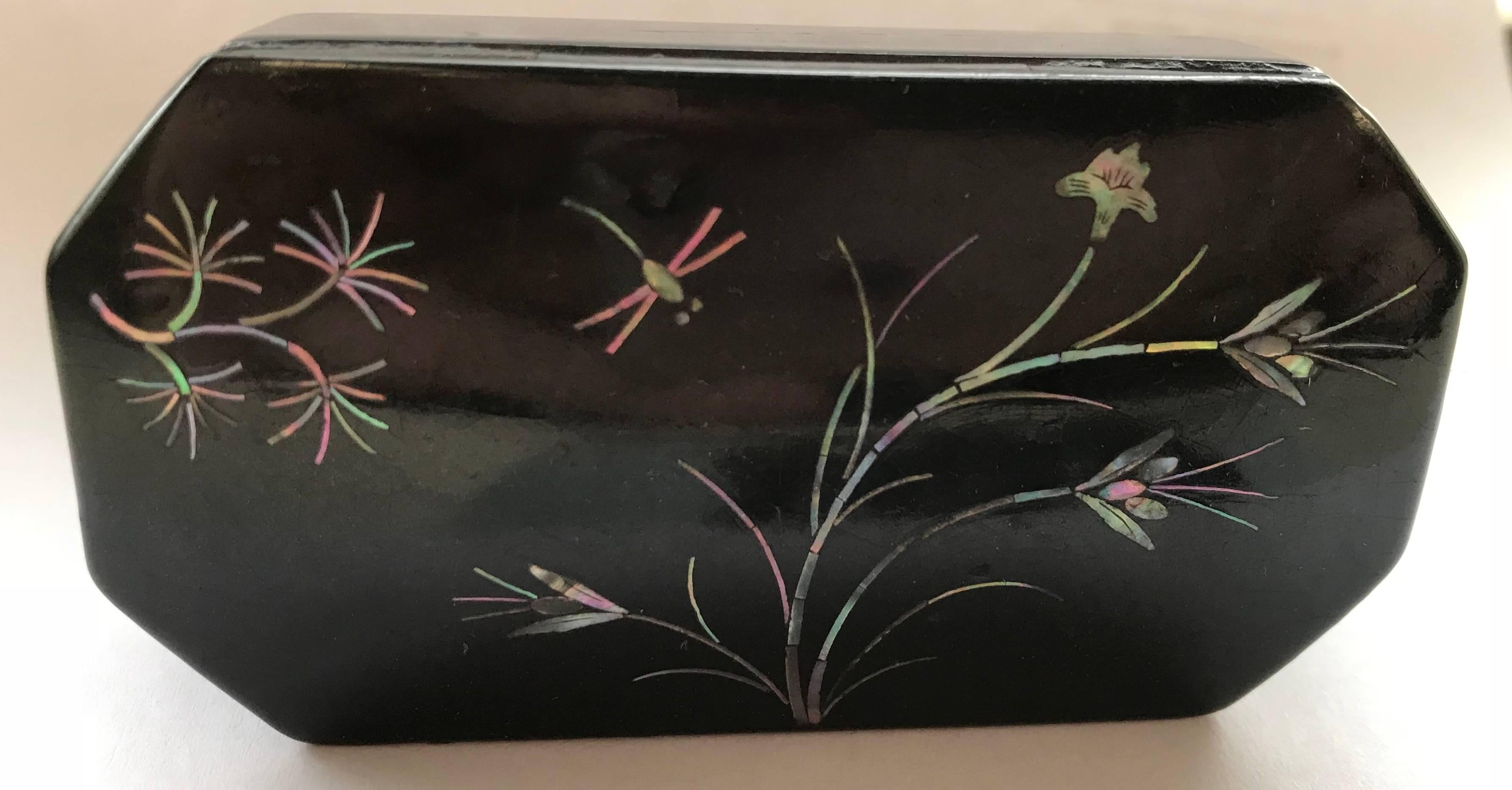 A dragonfly is flying amongst the inlaid mother of pearl flowers on this Asian black lacquer snuff box. It has angled lines to add to its character. The inside is lined in zinc.