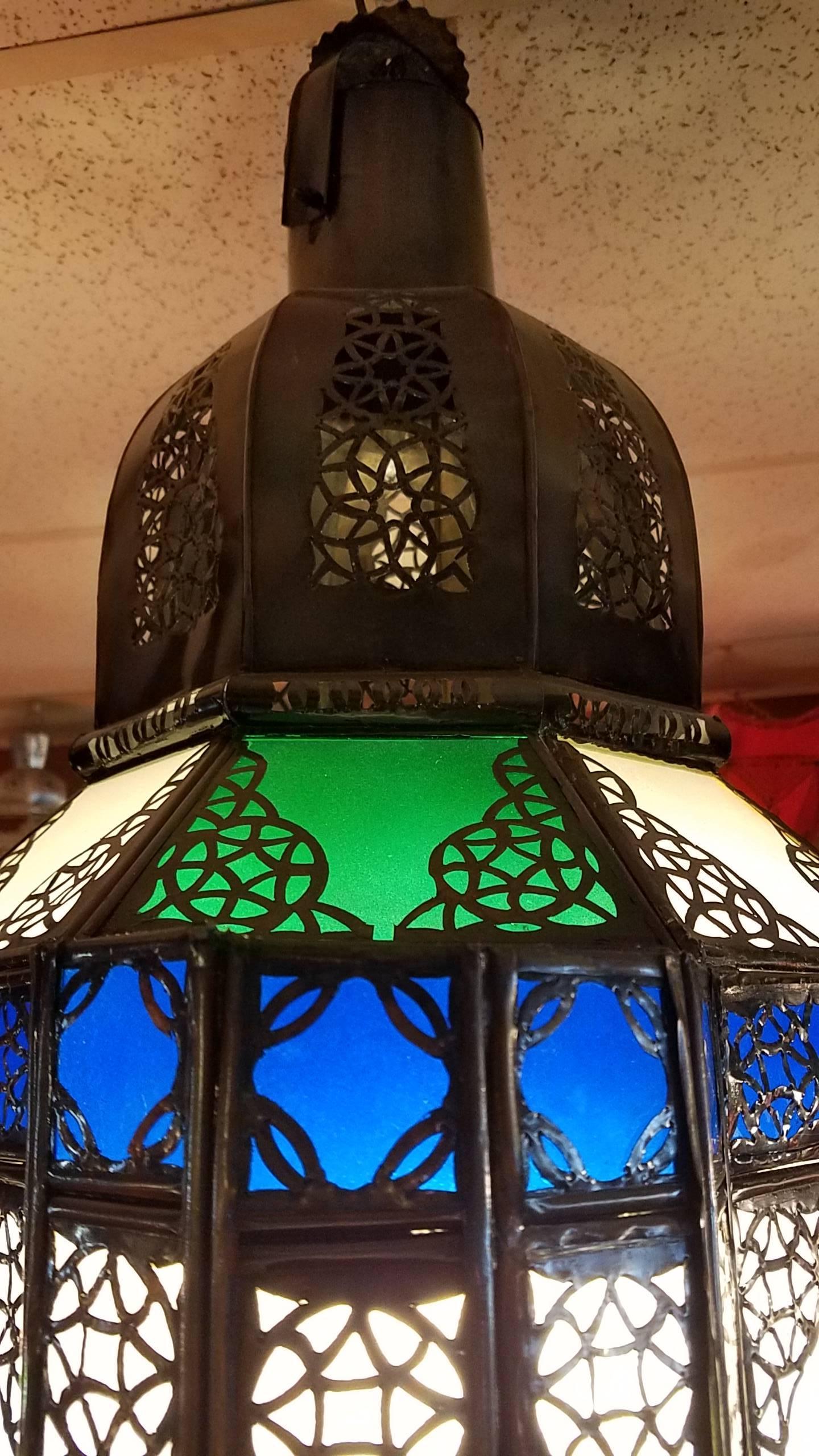 This is a beautiful Moroccan glass lantern, featuring frosty white glass panels, and many green, blue, and amber small glass patches. The metal frame is pierced throughout, to give the lantern an exquisite look from far as well as from up close.