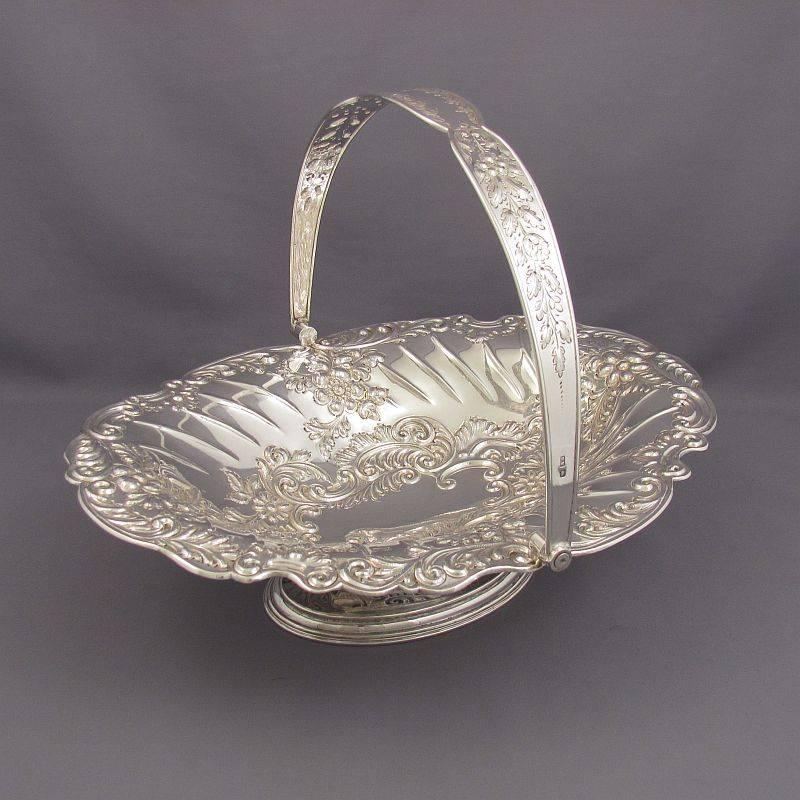 A Victorian sterling silver cake basket by Henry Wigfull, Sheffield 1896. Oval shape on pedestal foot with wing handle, decorated with repoussé flowers, foliage and scrolls. Measures: 12" (30.5cm) long, 19 ozt (592g).