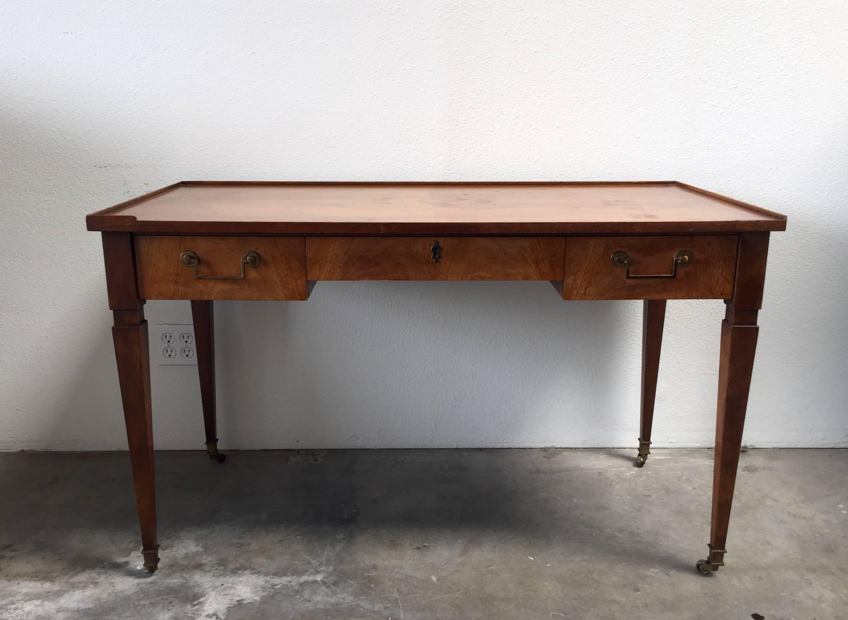 Elegant writing desk by Baker. Boasts three top drawers with brass hardware, stylist legs and brass wheels. Finished back allows this statement piece to float in any space. Makers mark in top right drawer.
