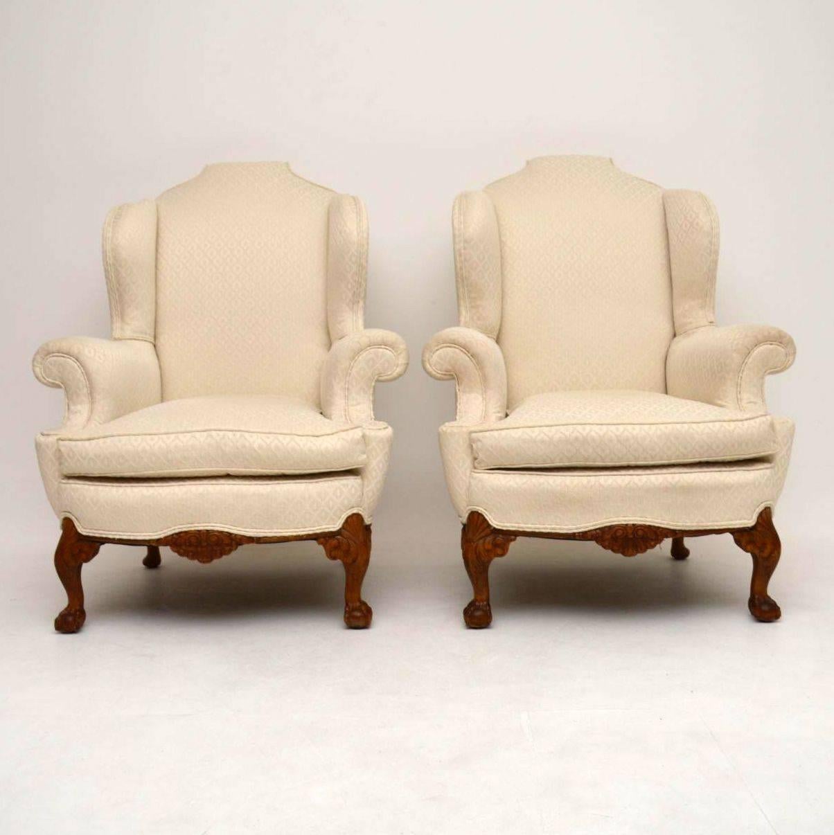 Very impressive and comfortable pair of antique wing armchairs in good condition and dating from circa 1920s period. They have high scroll over hump backs, wide wings and arms, loose cushions and sit on solid walnut carved frames. The carving on the