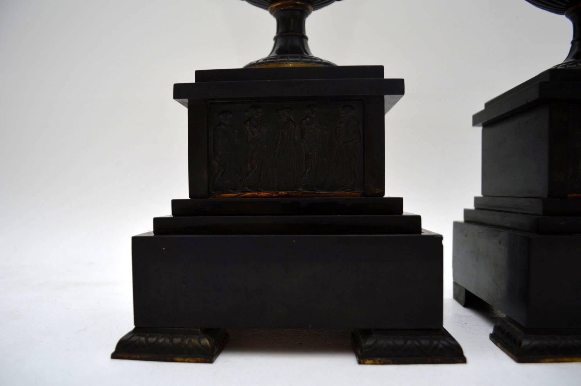 Pair of antique neoclassical bronze urns sitting on black marble bases in good original condition and with a nice patina. The urns themselves are embossed with warriors on chariots and the bases too have similar relief work on the fronts. These are