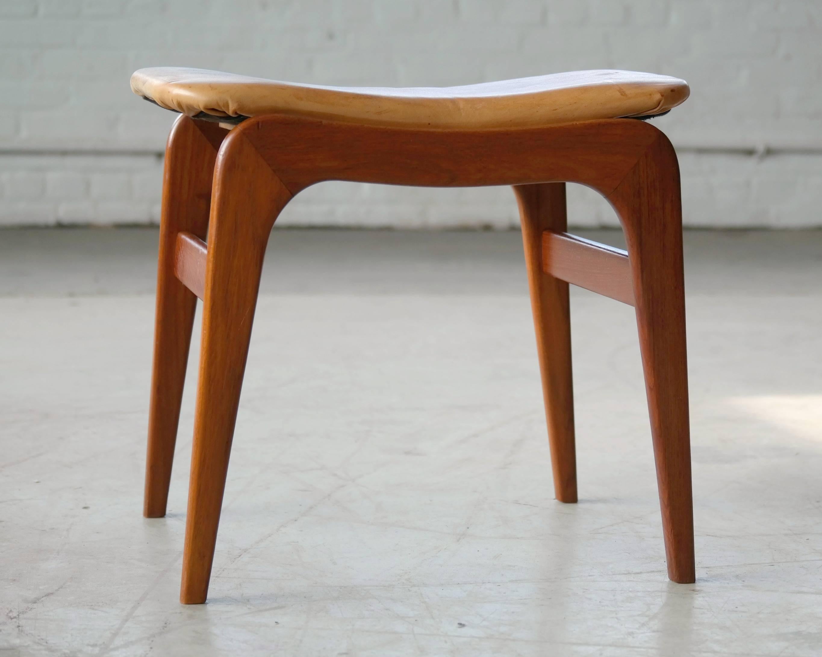 Danish Mid-Century footstool in teak and tan leather style of Fritz Hansen. The tan or cognac leather shows an admirable patina and a strong expression of wear, in the form of spots and marks which brightens up the natural leather.