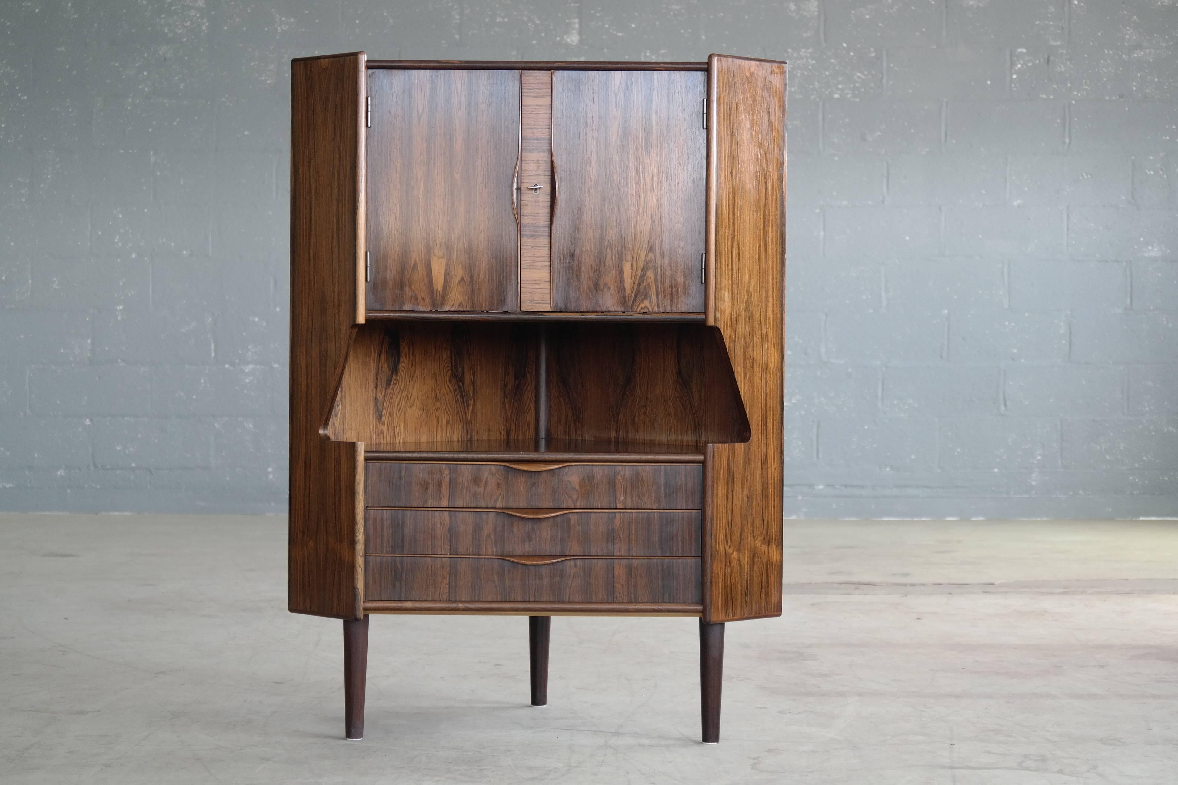 Fantastic corner cabinet and dry bar by Gunni Omann made in Rio Palisander with a beautiful deep color and grain. The top features a cocktail bar behind two doors with three storage drawers below. Great storage for bottles and glassware for home or