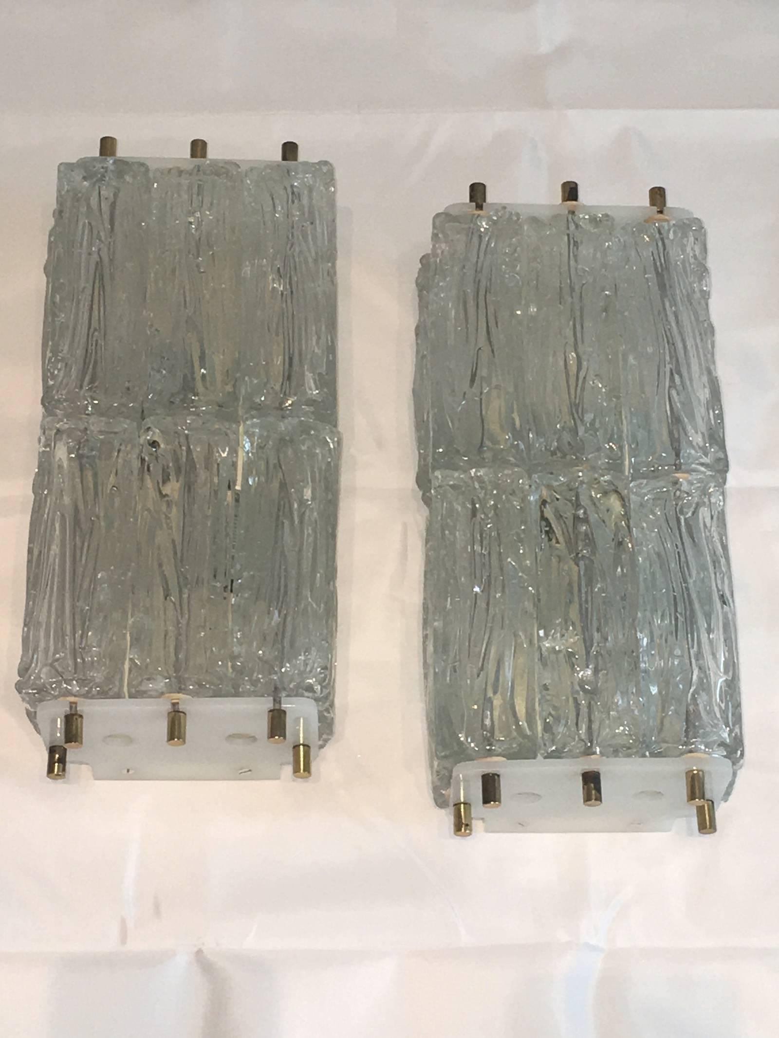 A pair of wall sconces, produced circa 1970s in the style of Venini, with rows of textured glass ice blocks as shades, each stack of three affixed to plastic mounts by brass heads, surrounding two sockets, against an enameled back plate. Overall