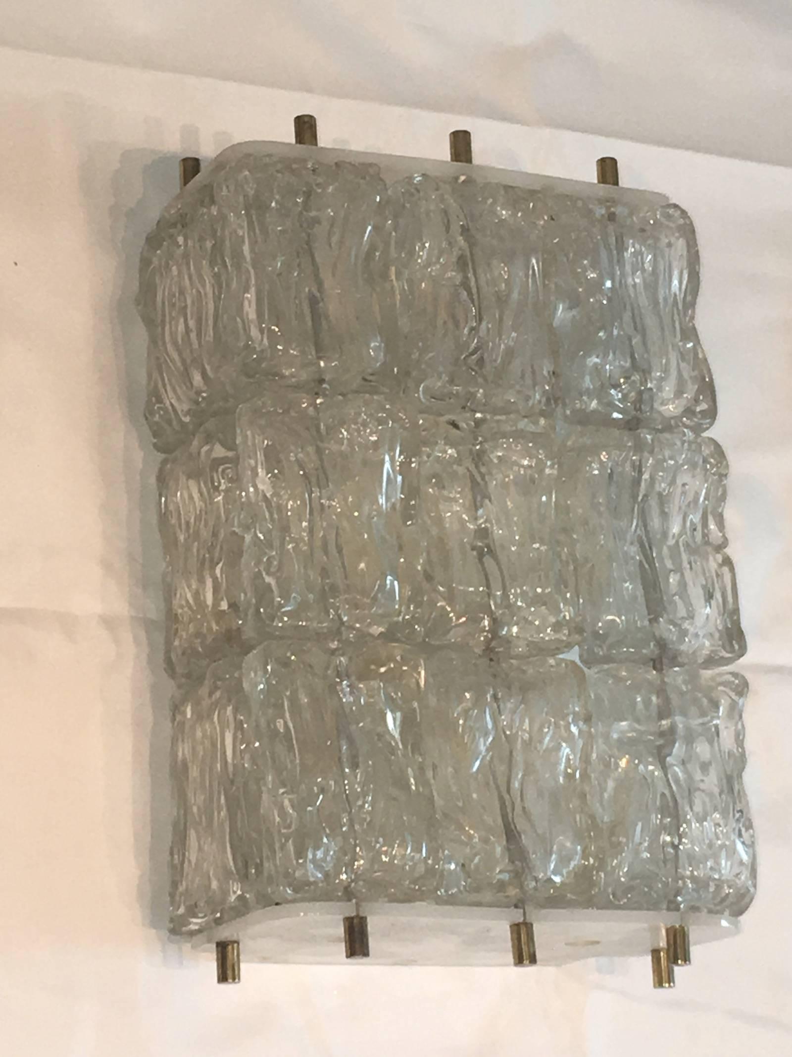 A beautiful wall sconces, produced circa 1970s in the style of Venini, with rows of textured glass ice blocks as shades, each stack of three affixed to plastic mounts by brass heads, surrounding four sockets, against an enameled back plate. Overall