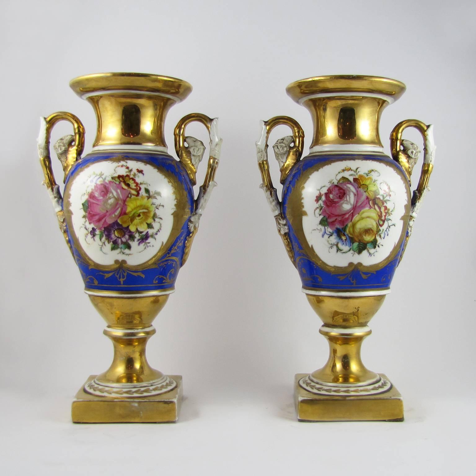 A beautiful pair of Amphora vases in white porcelain, gilded and cobalt blue painted.
The two handles in biscuit porcelain are transformed in ram heads on the upper end.
There is a polychrome decoration depicting floral subjects on both sides of