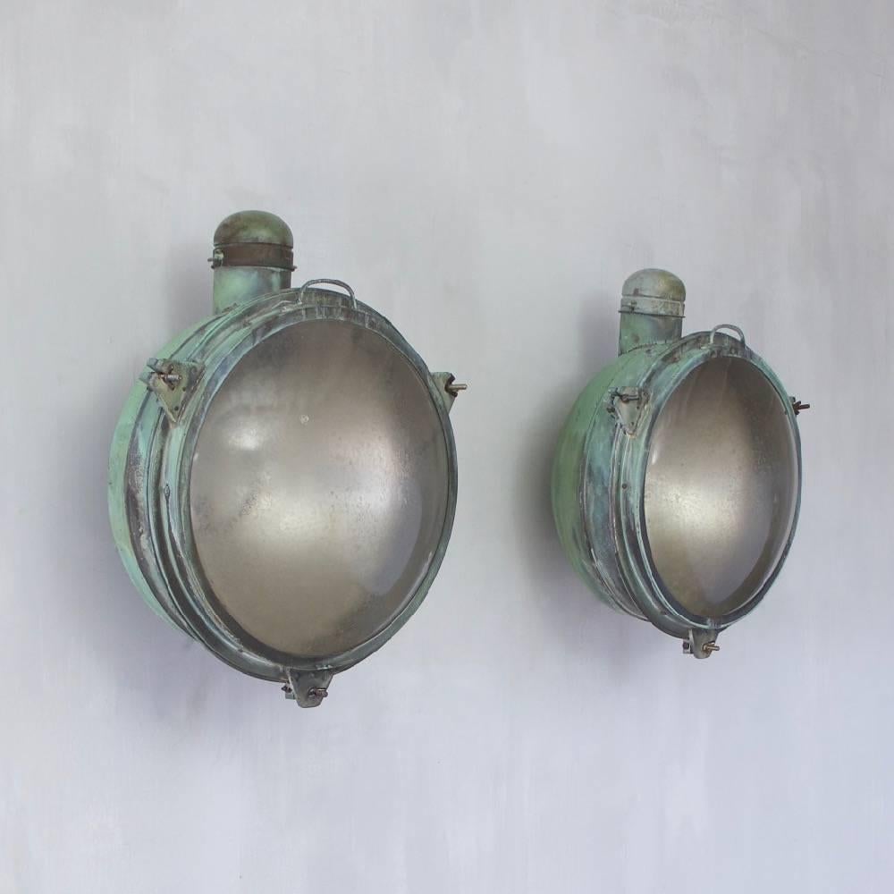 A very large pair of copper searchlights with hidden mounting brackets and original verdigris patination. The original glass shades stamped Holophane, England, circa 1920. One shade with a small chip to the surface - see images. Rewired with