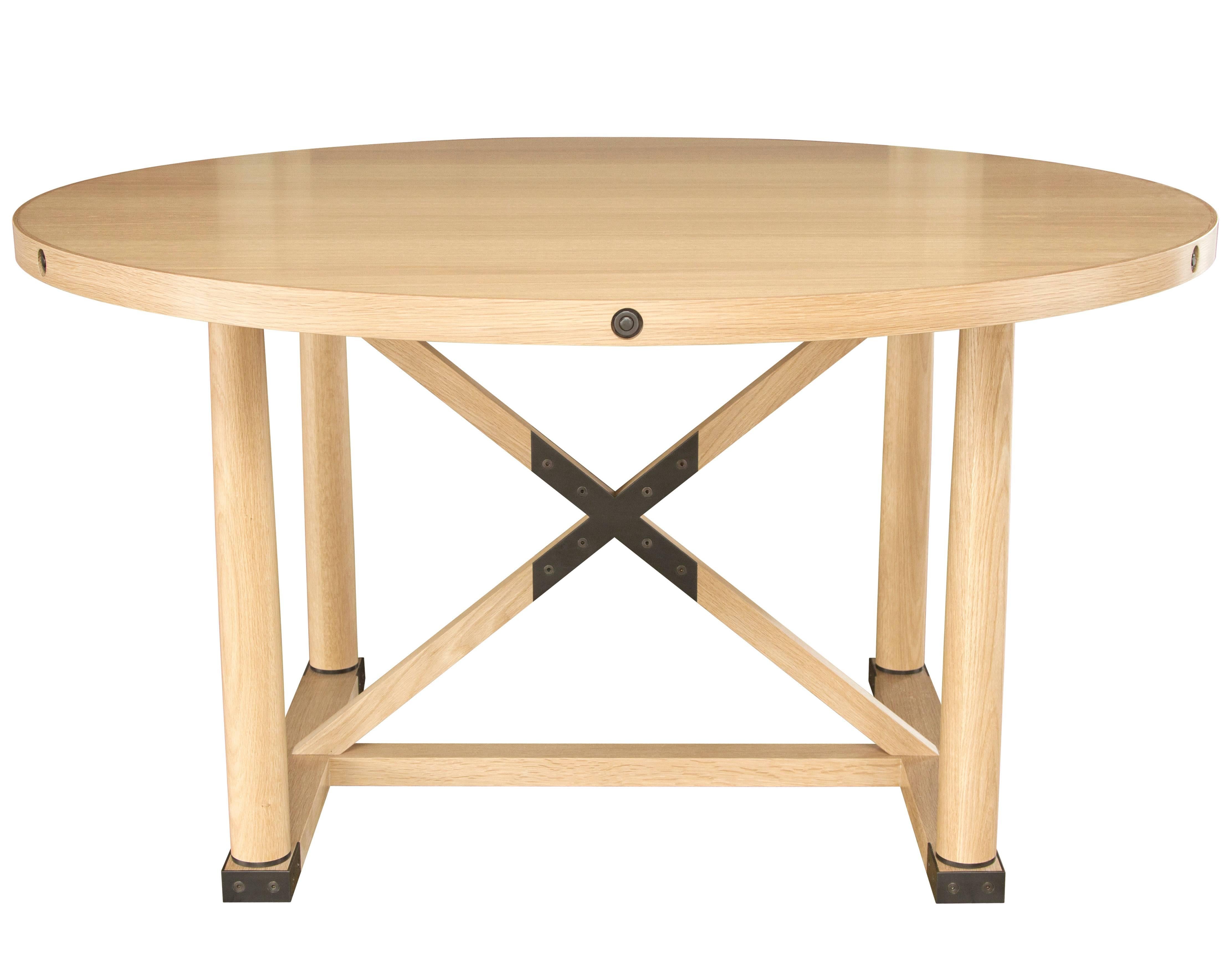 The Carden round table in natural finished white oak with darkened brass hardware. 
A striking silhouette featuring metal details in darkened brass. Four decorative side bolts around the table top edge, an inlayed central X brace, solid discs joined