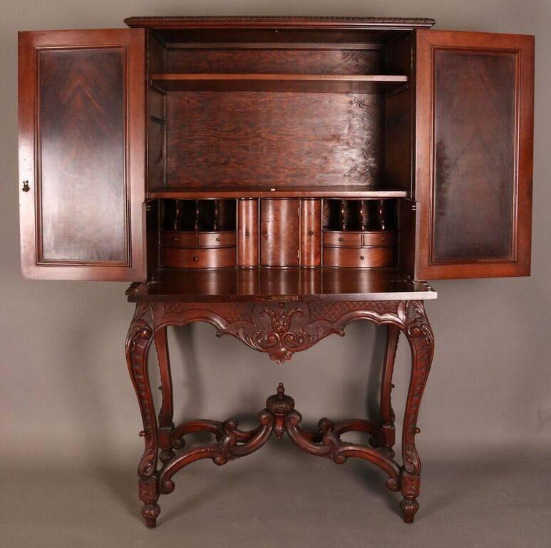 Antique Louis XIV style custom hand-carved French china cupboard features walnut construction with front door panels carved with festoons of flowers concealing drop front desk with drawers and storage compartments and scroll stretchers with central