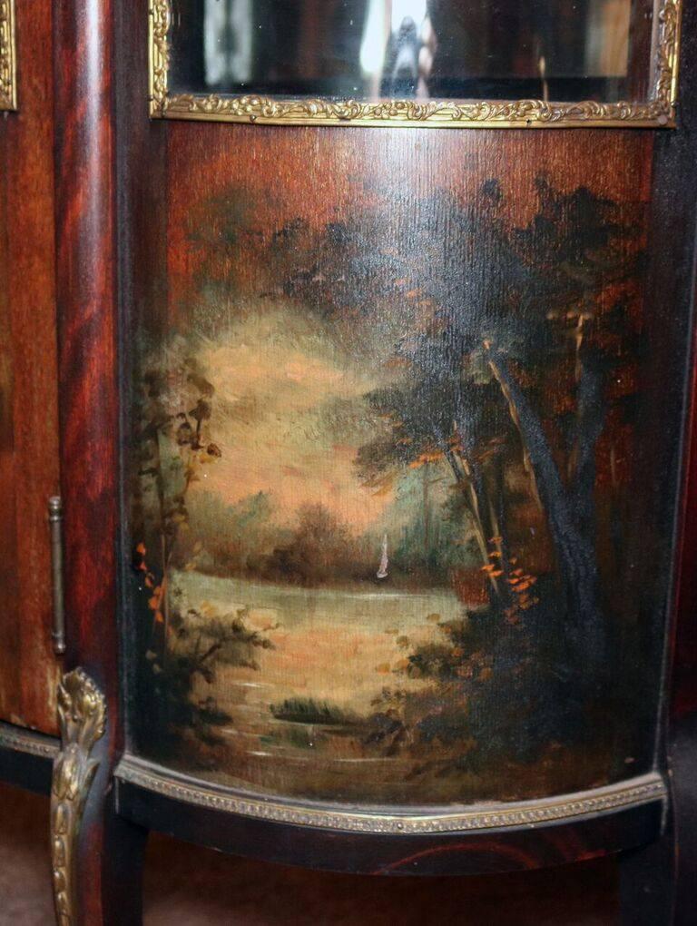 19th century French Louis XV style vitrine features mahogany construction with hand-painted vernis martin courting and landscape or lake scenes, traditional cabriole legs, ormolu accoutrements, bronze gallery atop, and curved glass sides and door