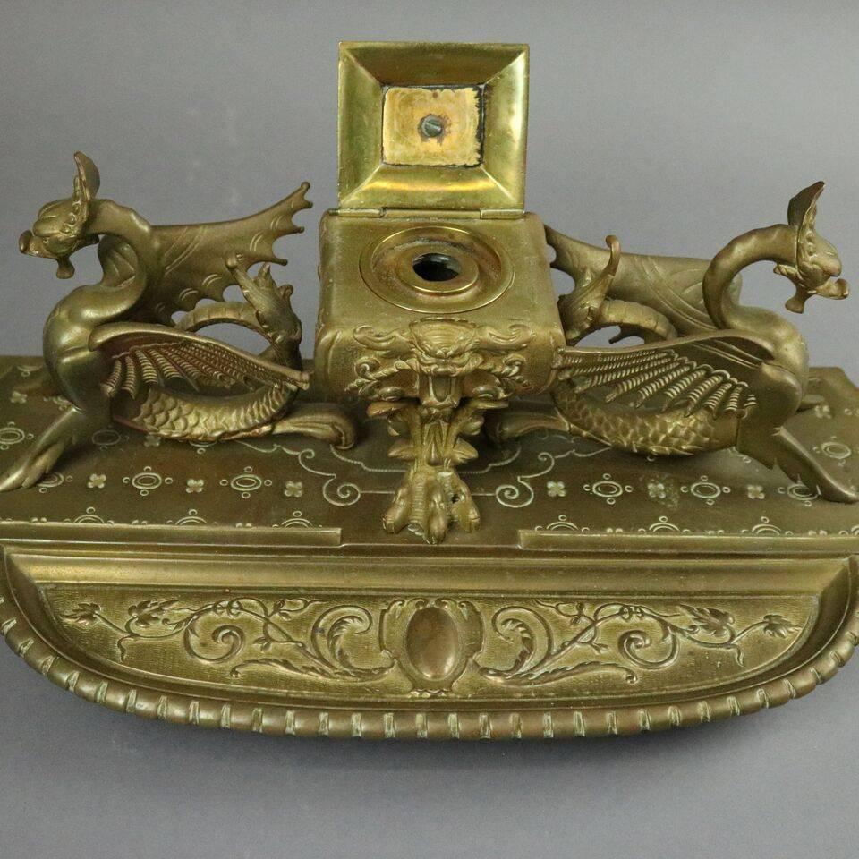 Antique figural bronze Egyptian Revival inkwell features raised inkwell with central mask and hinged crested lid flanked by opposing sea dragons on footed base with pen tray of scroll and foliate design, late 19th century.