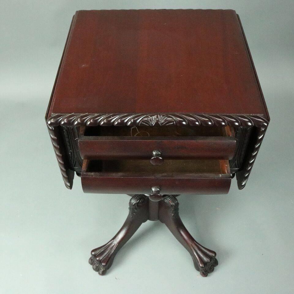 Antique American Empire stand features mahogany construction with drop-leaf top bordered by carved twisted rope atop a case with two-drawer flanked by carved foliate panels with corner drop finials supported by acanthus leaf shaft on plinth