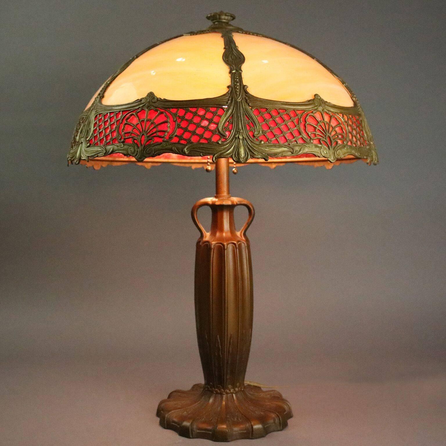 Antique Arts and Crafts Royal Lamp Co. table lamp features six-panel shade with cream and red slag glass in a foliate filigree bronze frame which covers two lights supported by a double handle bronze base, newly re-wired, circa 1920

Measures: