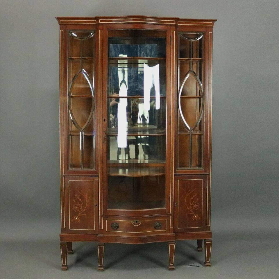 Antique mahogany Edwardian mirror-back china cabinet features centre serpentine glass door flanked by leaded bevelled glass panels above blind storage compartments and drawer, doors decorate with floral inlay and mother-of-pearl accents, banding