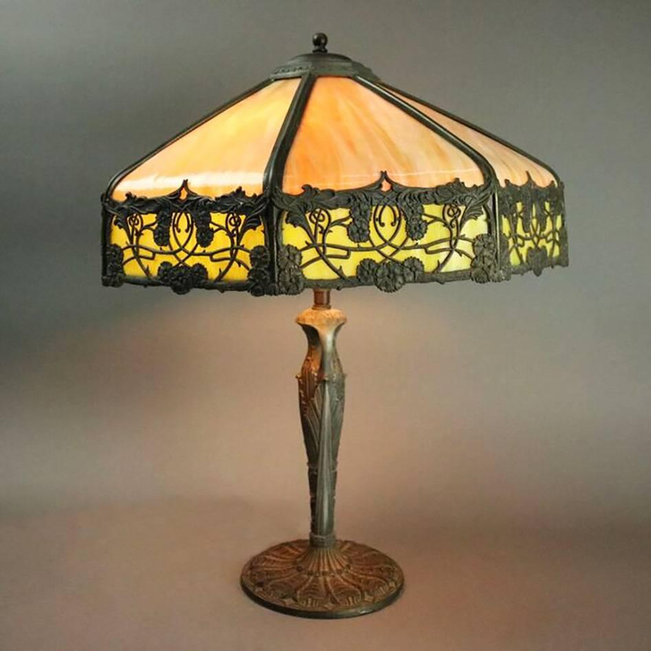 Antique Arts and Crafts Bradley and Hubbard School dual light table lamp features foliate filigree bronze eight panel shade with caramel and green slag glass seated on cast double handle white metal base foliate decorated with acanthus, re-wired,