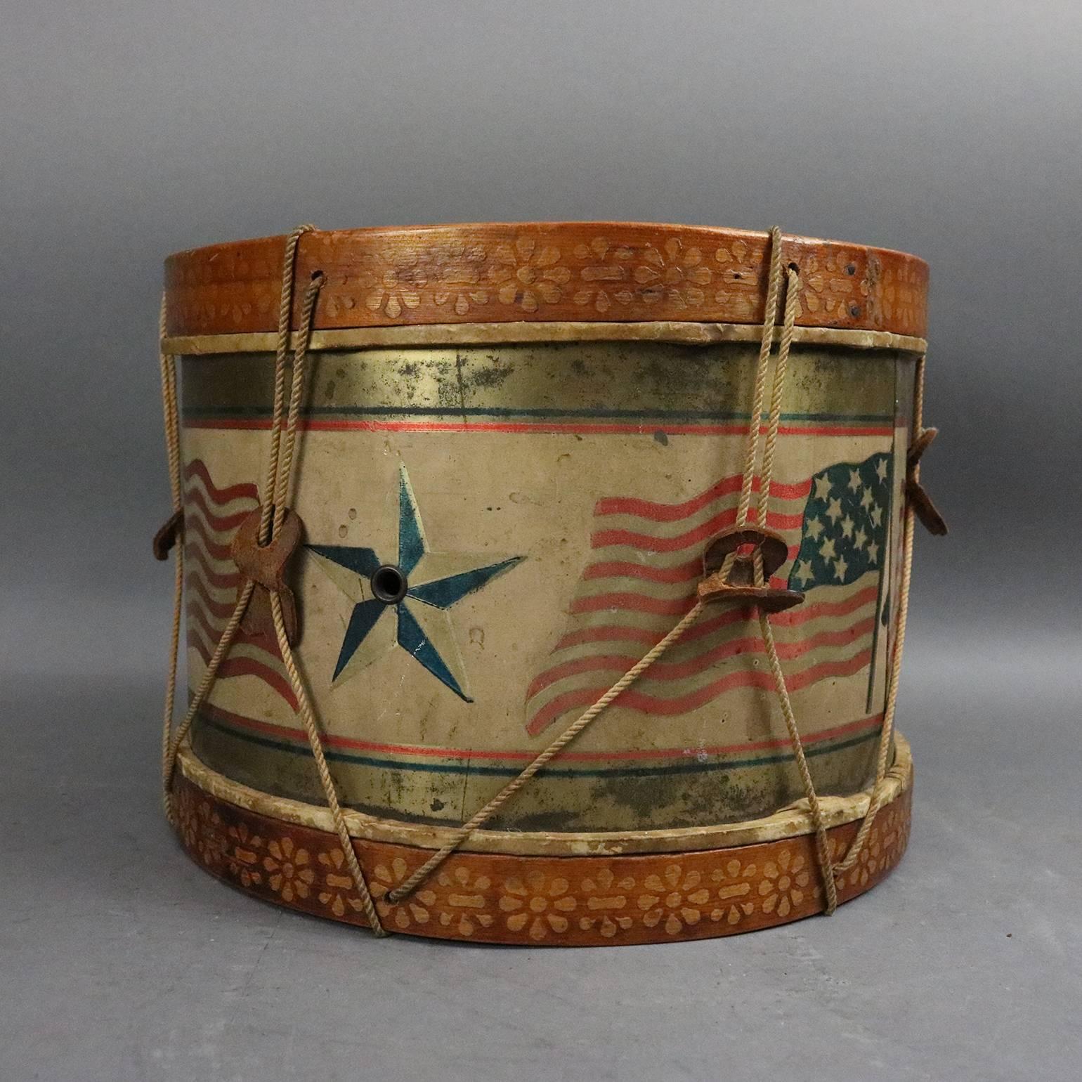 Americana fold art tin drum features patriotic theme of 13-star American flags and stars Stenciled around mid-portion, bordered by Stenciled floral motif upper and lower, leather drum head, circa 1900.

Measures: 8" Height x 11" Diameter