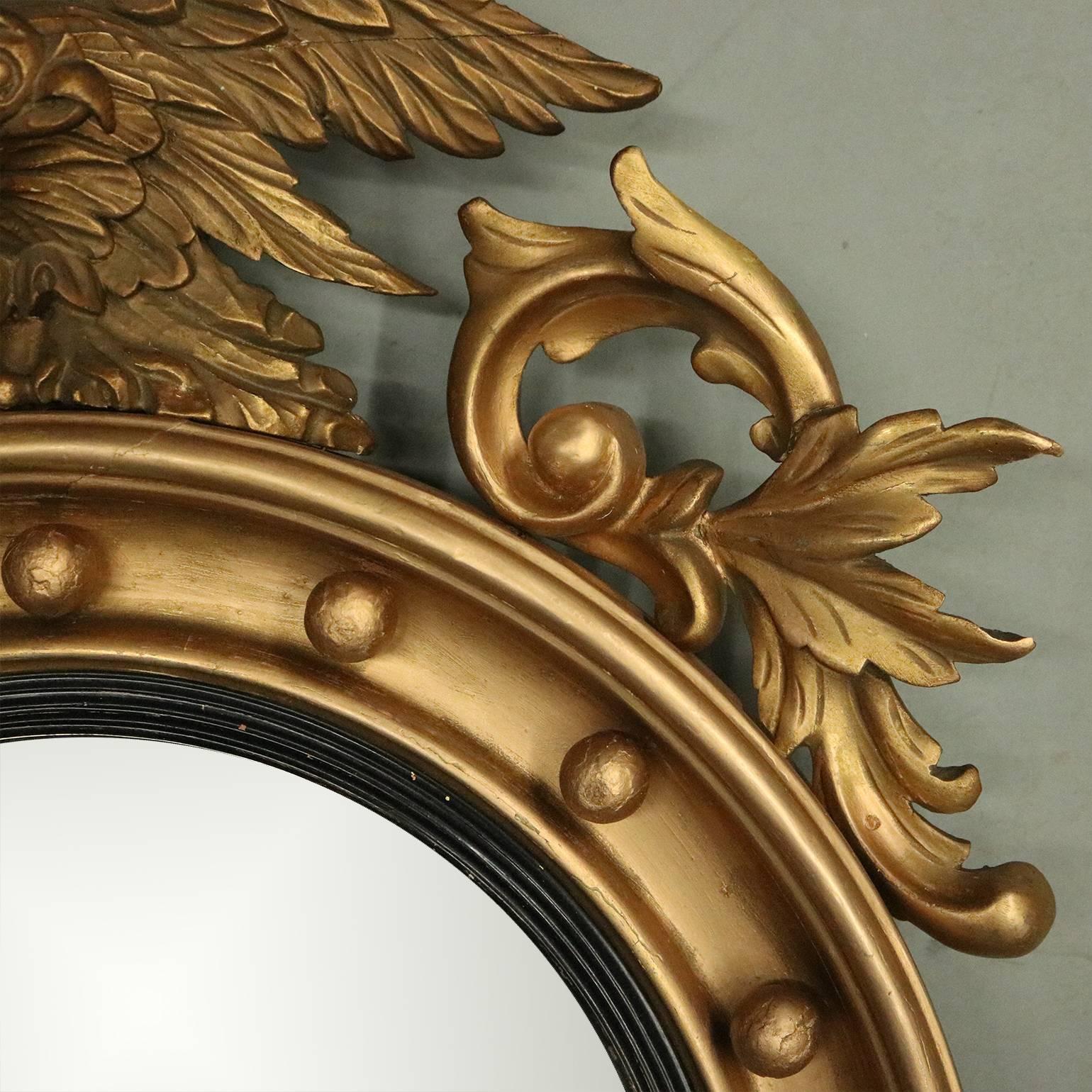 Antique Federal bullseye mirror features the original convex plate bordered by an ebonized fillet within a gilt gesso frame interspaced with 16 spherules, surmounted by an eagle atop a rocky mound and flanked by scrolled acanthus, circa