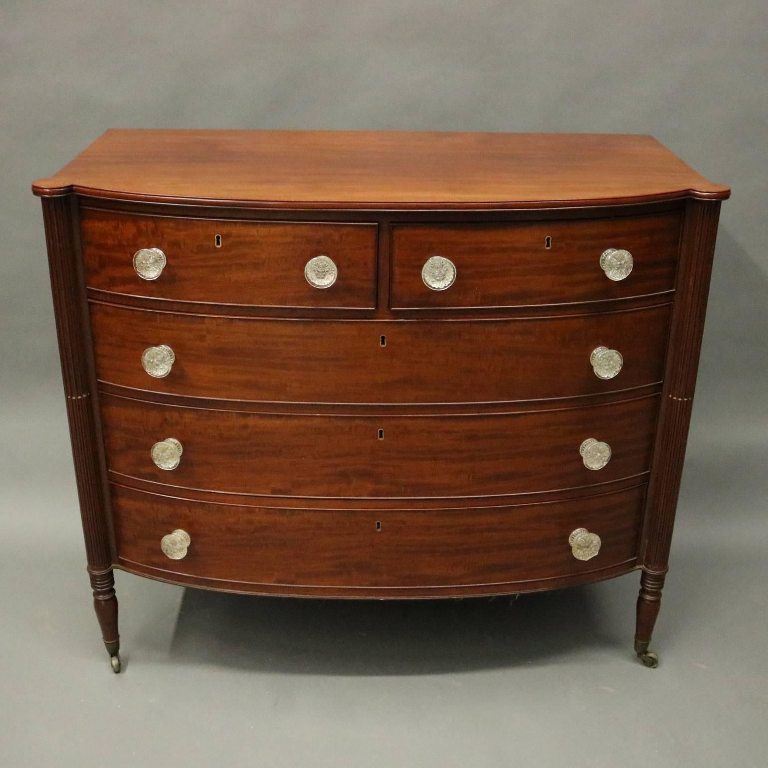 Antique Sheraton bow front chest of drawers features mahogany construction, cabinet with three locking long and two locking smaller upper drawers, one segmented for silver, flanking reeded columns, seated on tapered turned legs, molded glass pulls,
