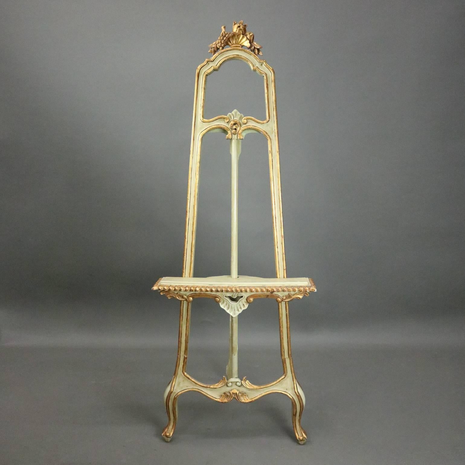 Vintage French, Louis XIV style adjustable art easel features gilt decorated painted wood construction with foliate, bead, and shell decoration, seated on cabriole legs and topped with carved fruit and nut pierced crest, adjustable, circa