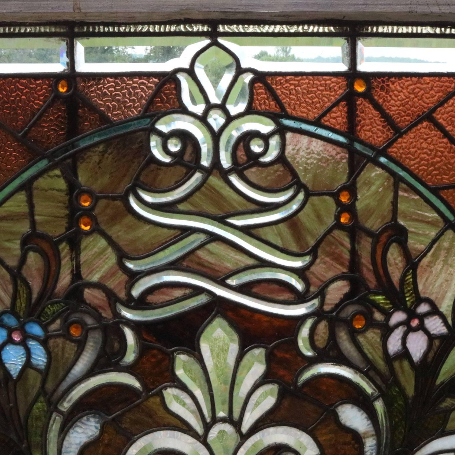 Antique Art Nouveau leaded stained glass window features leaded stained and slag glass panels in foliate and floral decoration with jeweled accents and lower central beveled glass design reminiscent of fluer-de-lis, bordered by leaded beveled glass