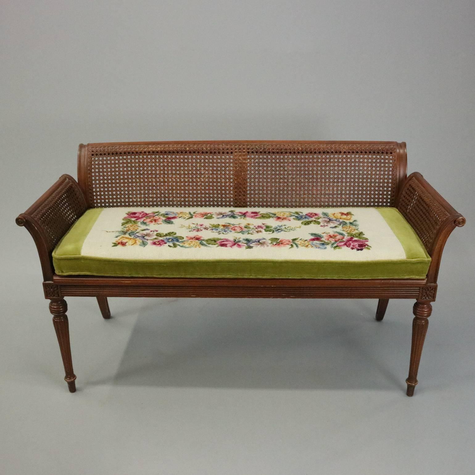 Antique French carved mahogany bench settee features caned seats, back and arms seated atop reeded framed with carved floral die joints over fluted and reeded legs and topped with floral needlepoint tapestry and velvet cushion, circa