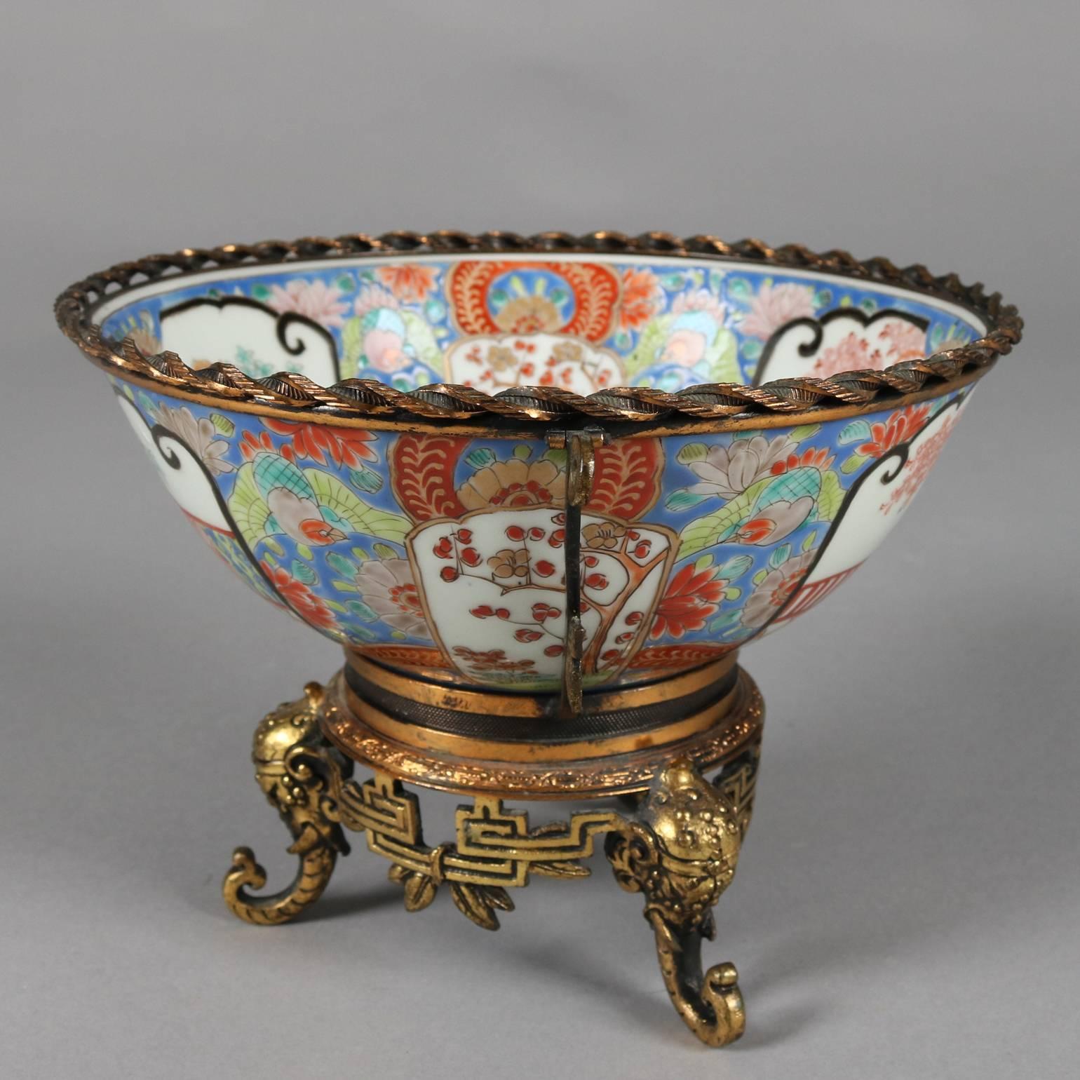 Antique Japanese porcelain Imari bowl features hand-painted porcelain with floral and foliate design with reserves depicting garden scene of courting couple, figural foliate and elephant cast bronzed footed base and twisted double handle frame,