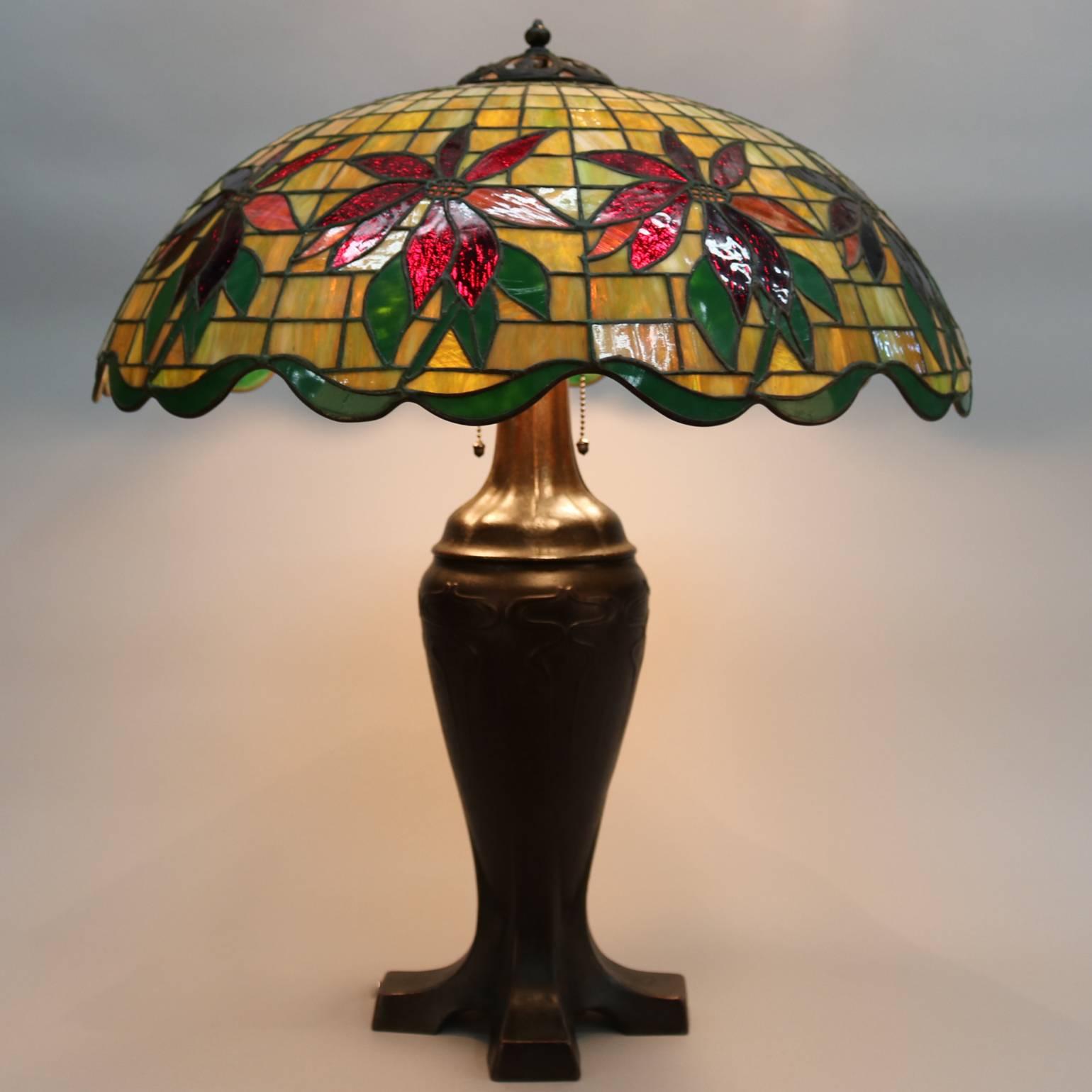 Antique handel leaded stained glass lamp features dome shade with mosaic poinsettia design, the footed standard with an Art Deco form rising to support a three-socket cluster, reminiscent of lamps by Louis Comfort Tiffany of Tiffany Studios, circa