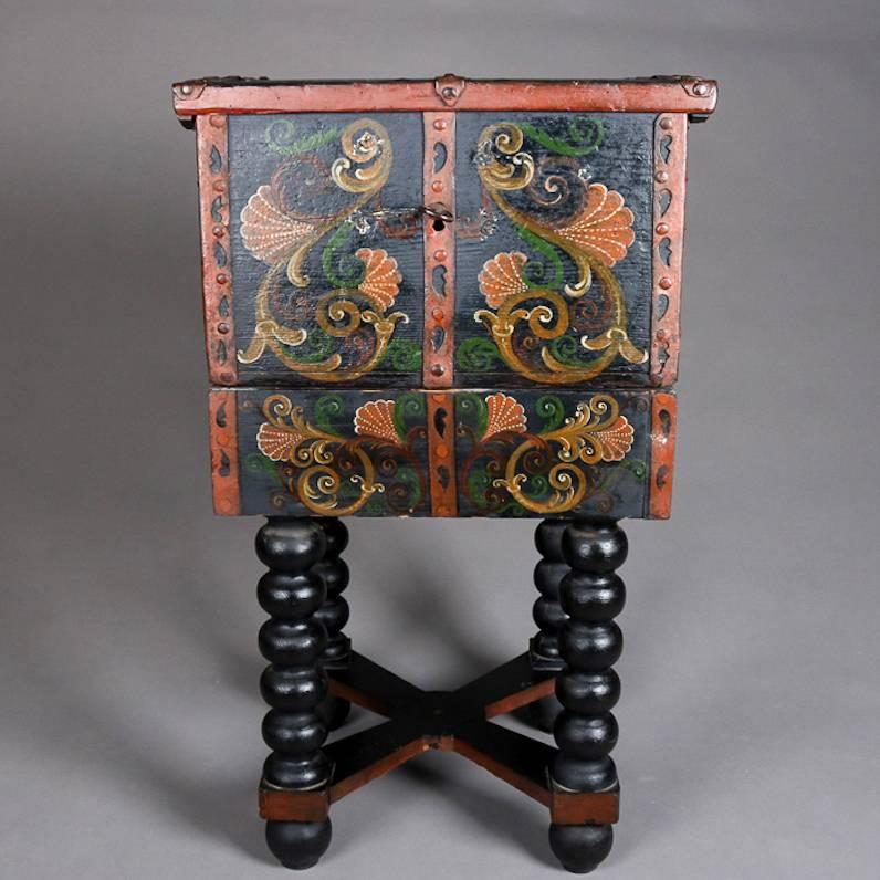 Antique Spanish Baroque chest features hand-painted scroll and foliate decoration on ebonized box with hand-wrought banding and bronze handles, seated on matching stand with turned legs and X-form cross stretcher, 19th century

Measures - 32"